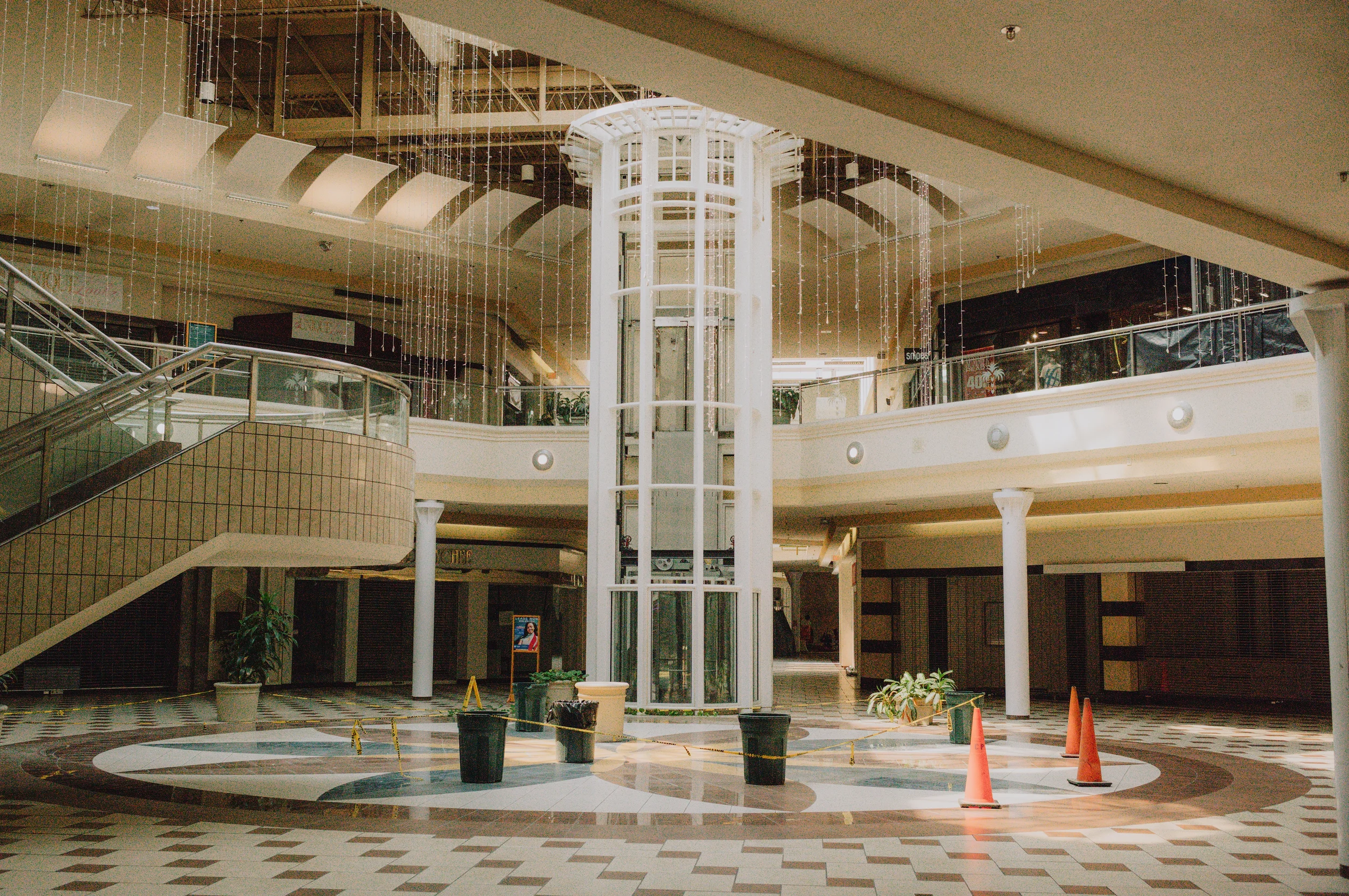 Interior of a shopping mall with a vacant elevator shaft and construction cones, potted plants, and a staircase on the left.