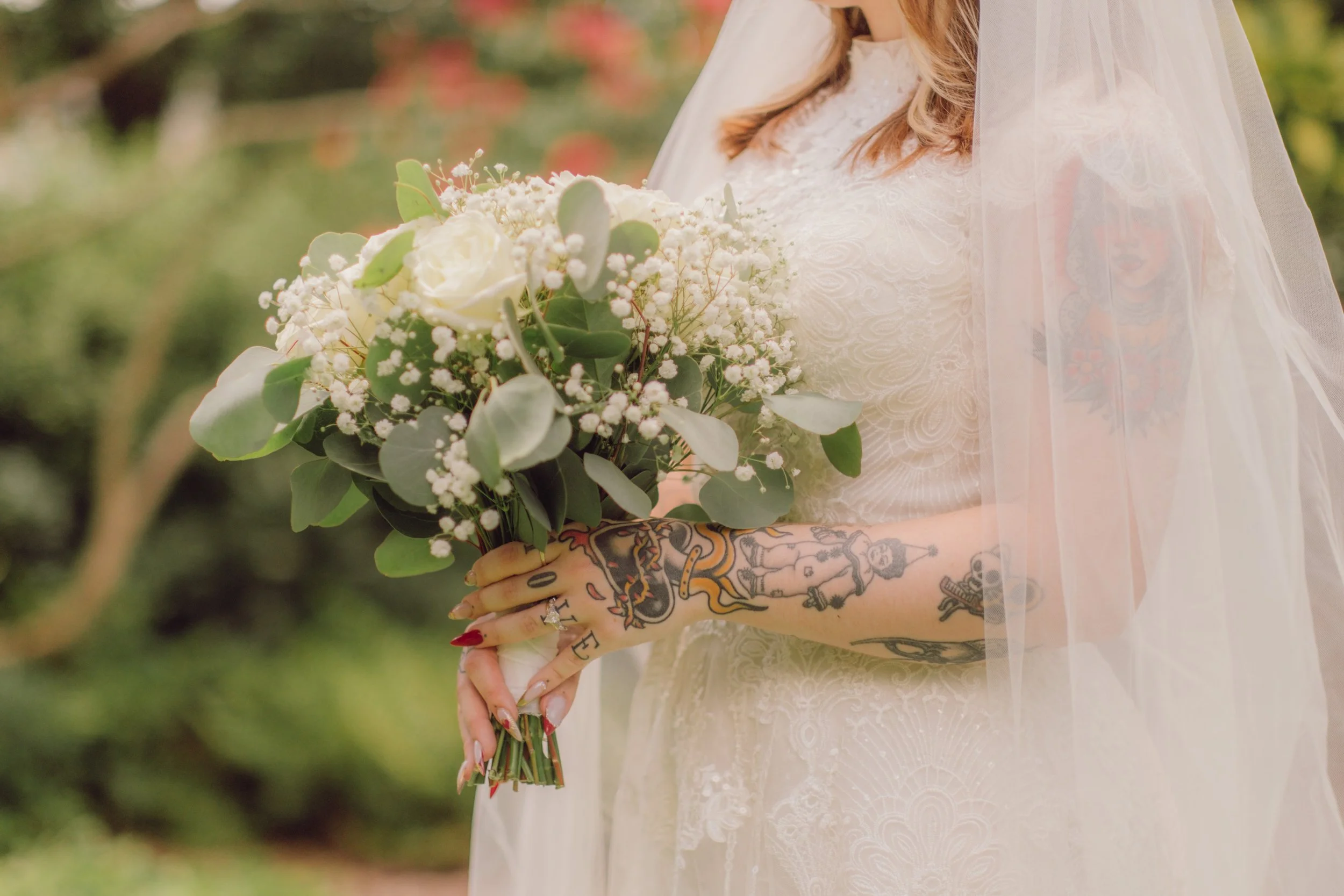 A bride holding a bouquet of white roses and greenery, with tattoos on her arms and a lace wedding dress, outdoors.