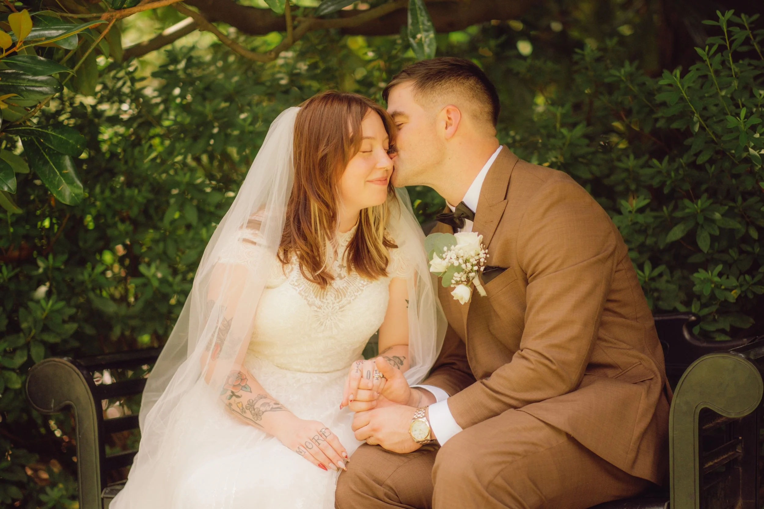 A newlywed couple sitting on a bench outdoors, with the groom kissing the bride on the forehead, both smiling softly. The bride wears a white wedding dress and veil, has colorful tattoos on her arms, and holds hands with the groom. The groom wears a