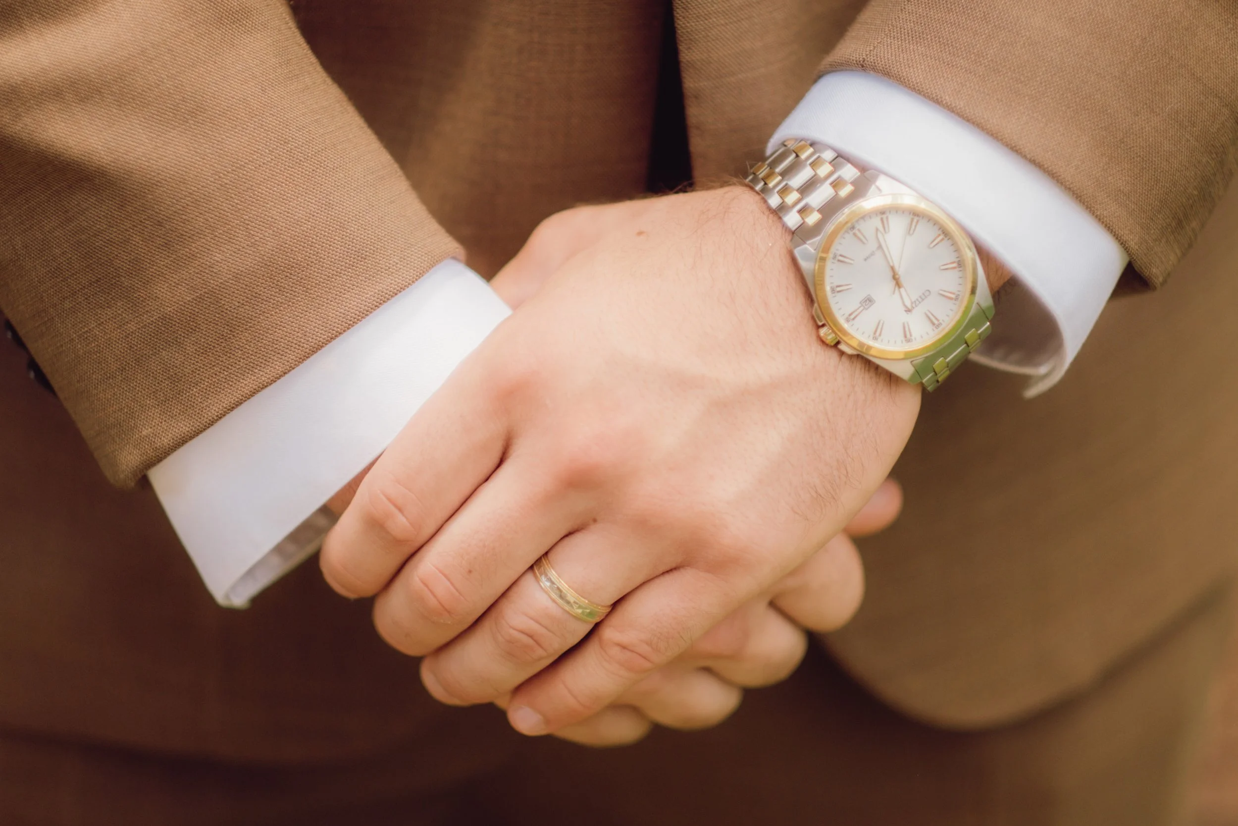 A person wearing a brown suit with white shirt cuffs, showing a gold and silver watch and a wedding band, with hands clasped.