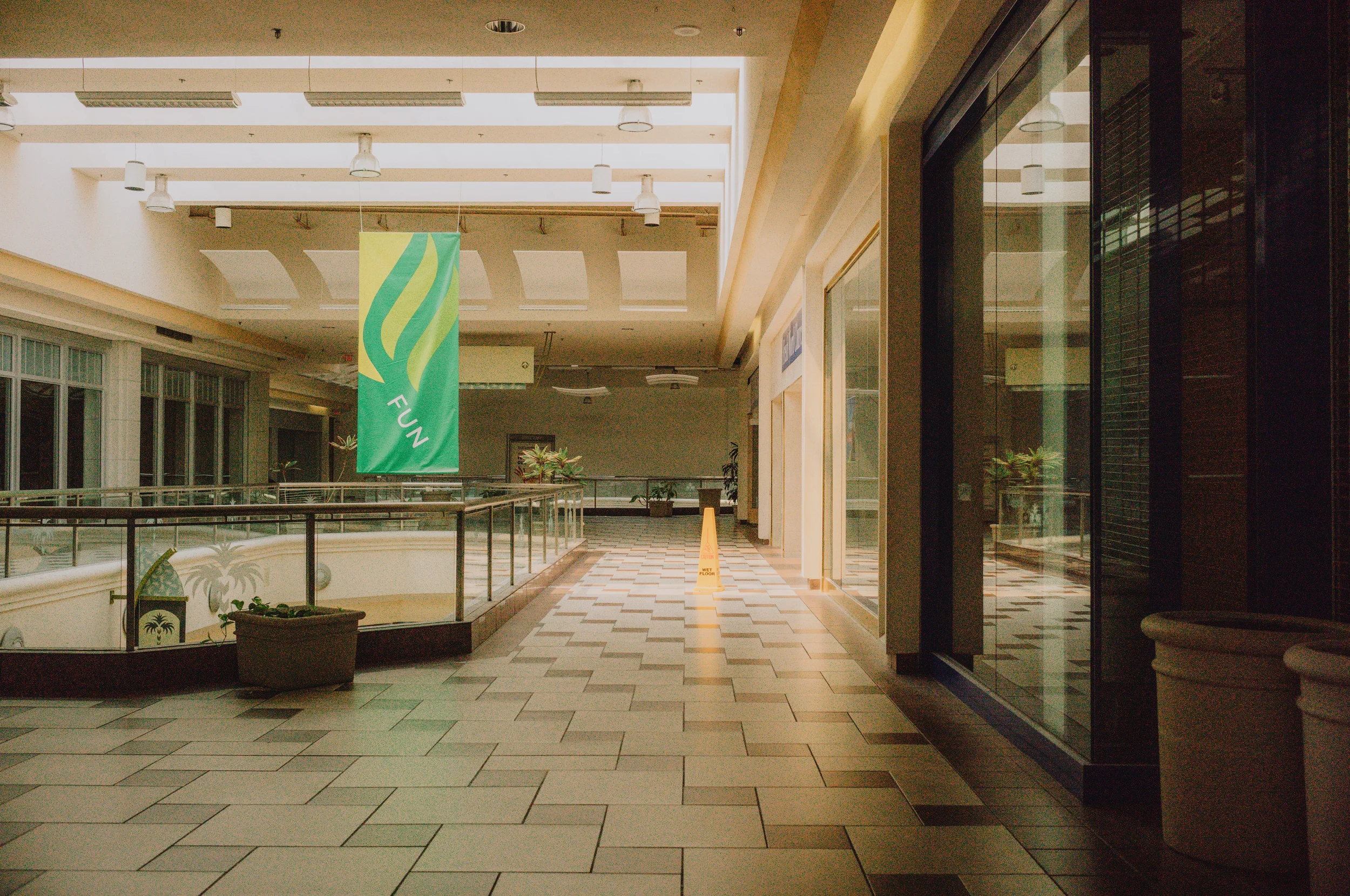 Empty indoor shopping mall corridor with tiled floor, potted plants, glass storefronts, and hanging lights. A green banner with the word 'FUN' hangs from the ceiling, and a yellow warning sign is on the floor. The scene appears quiet and unoccupied.
