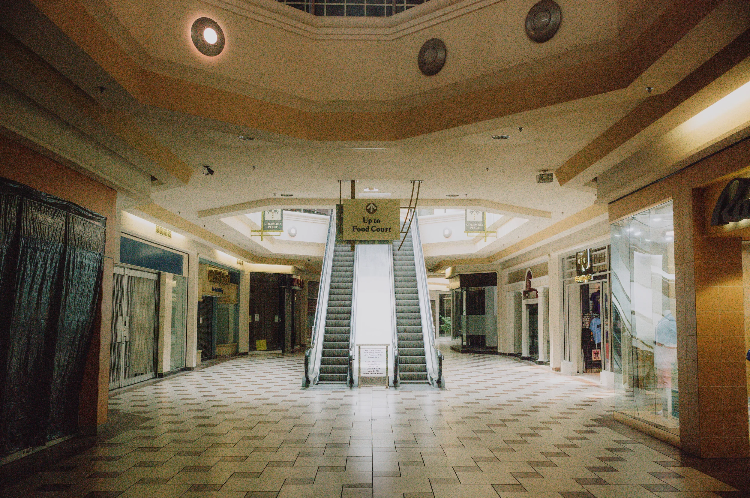 Empty shopping mall corridor with an escalator leading up to a food court, flanked by shops and stores on both sides.