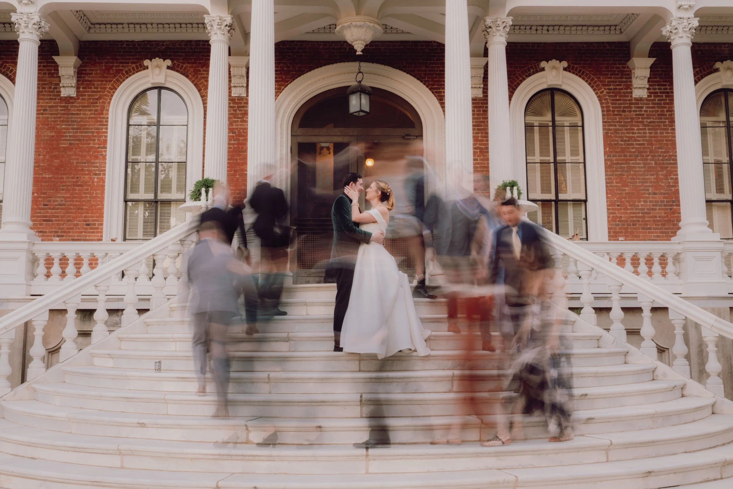 A bride and groom sharing a kiss on the steps of a historic building, with many blurred wedding guests moving around them.