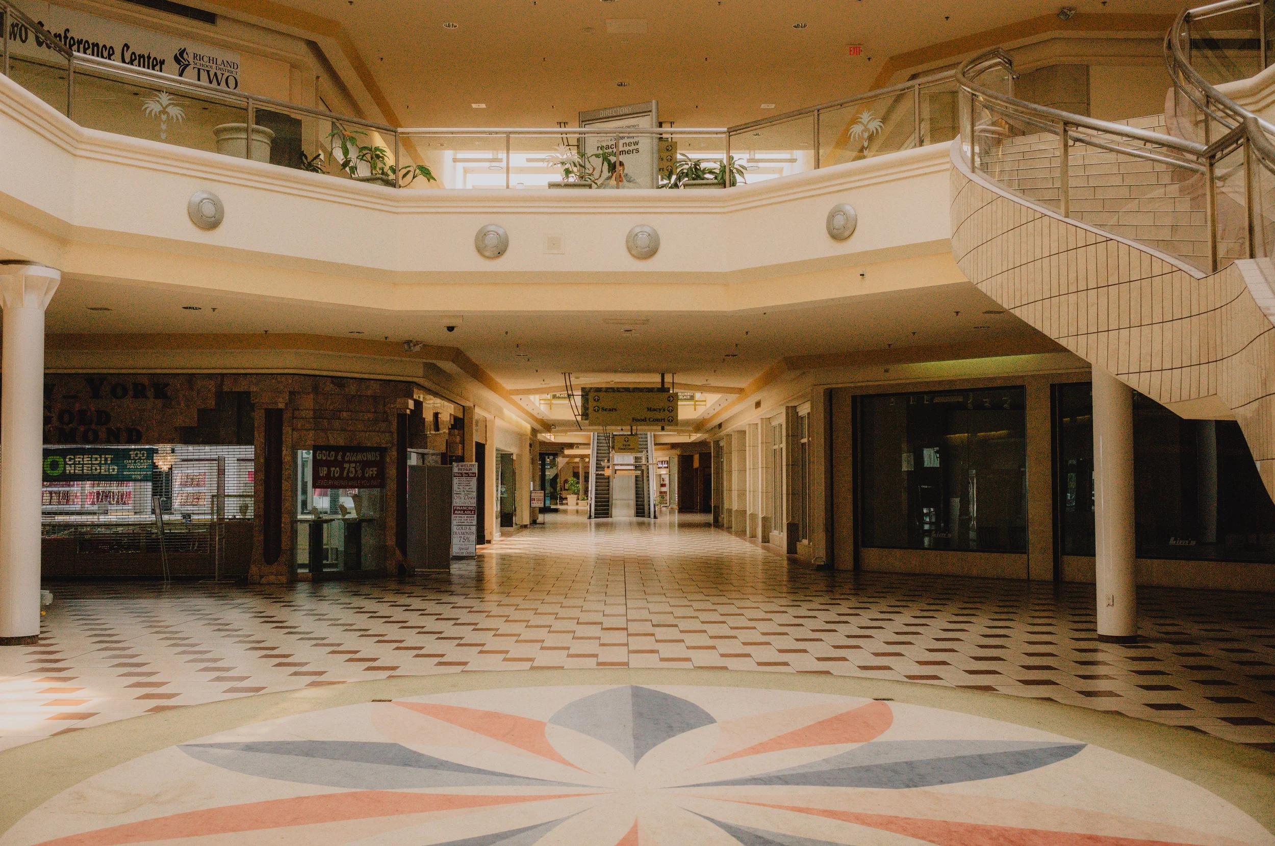 Interior of an empty shopping mall with multiple Levels, escalators, and storefronts visible. The floor has a geometric pattern, and there are signs hanging from the ceiling indicating directions to stores like Sears, Macy's, and Food Court.