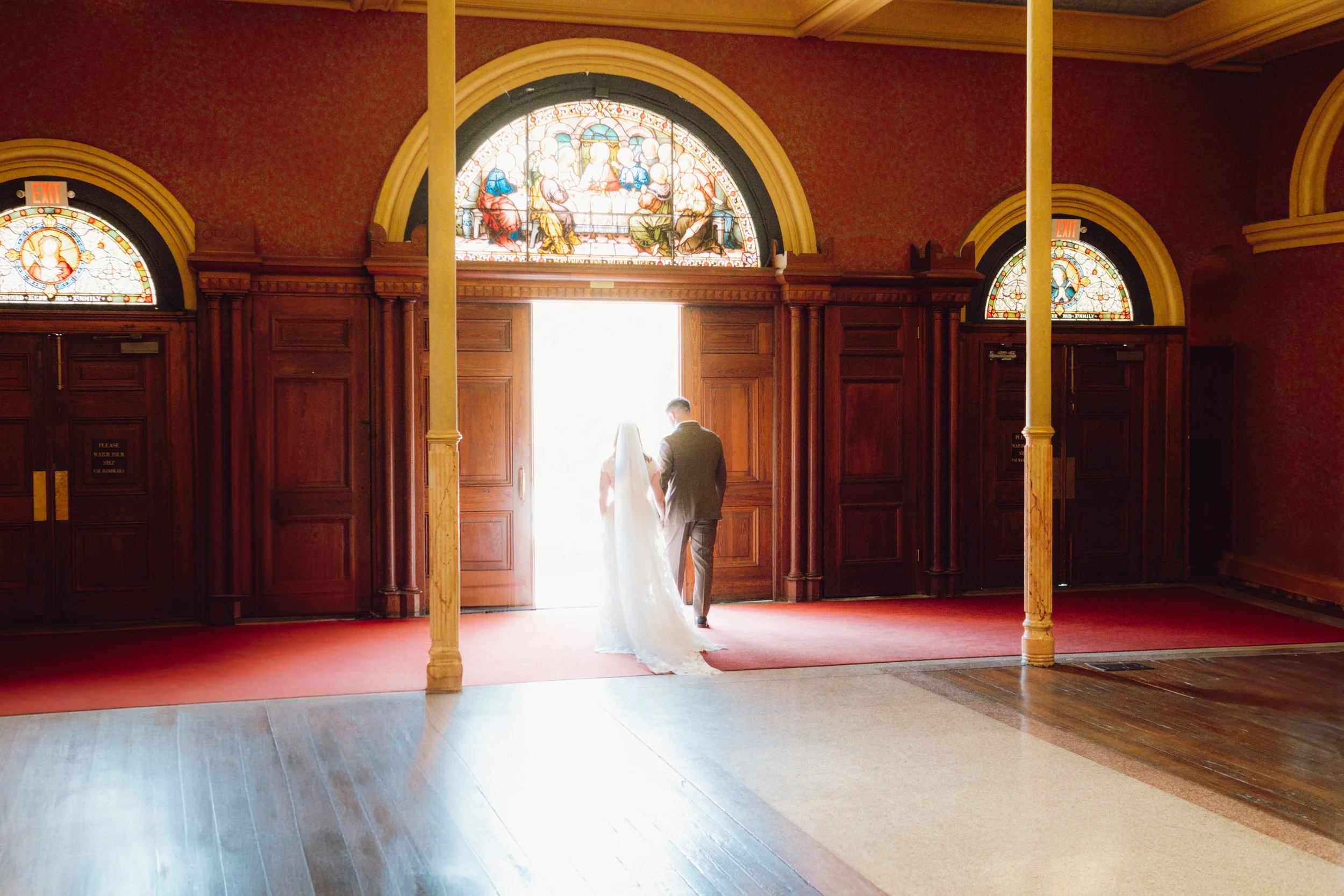 A bride and groom holding hands as they walk through an open door into bright light inside a church with stained glass windows and wooden paneling.
