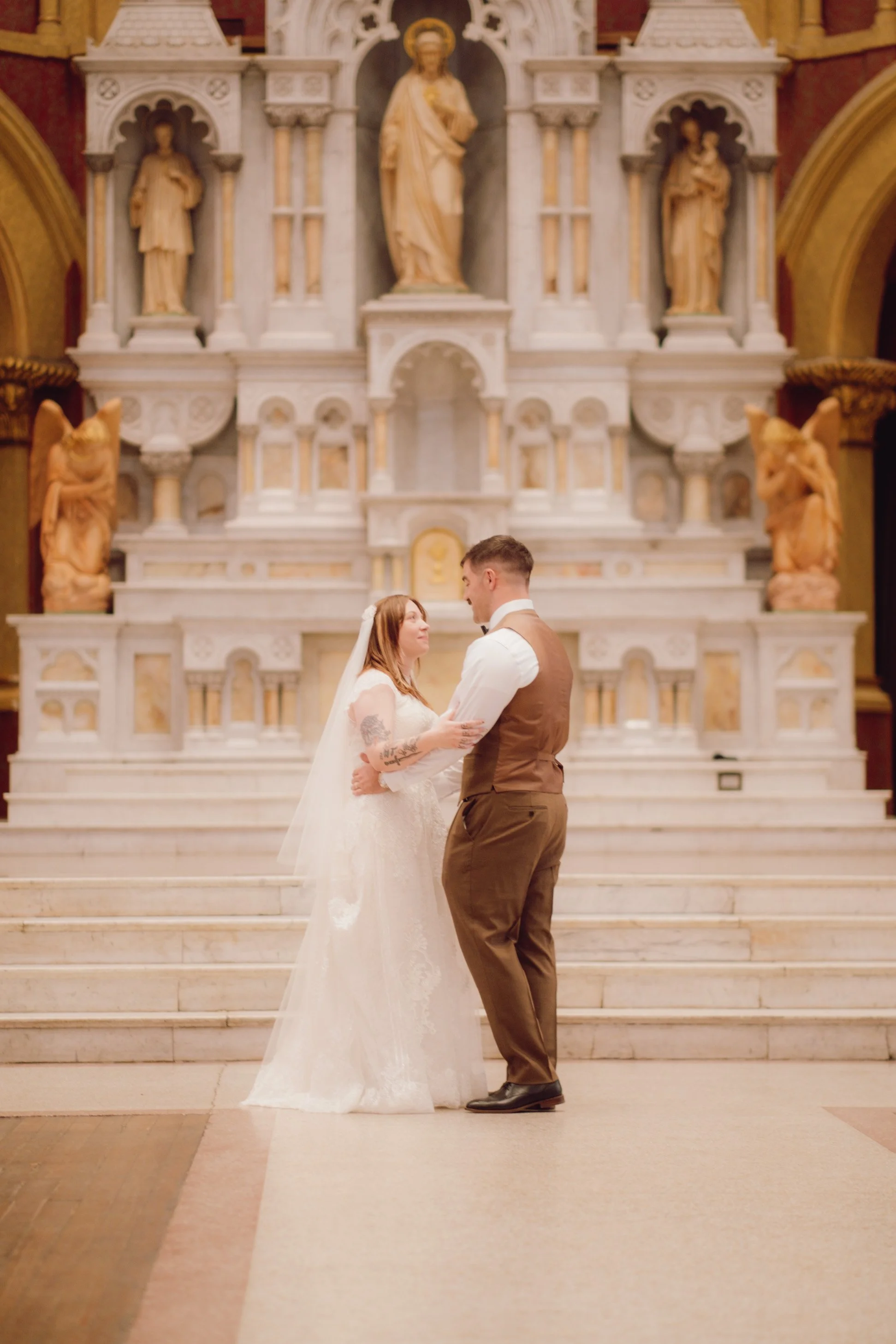 A bride and groom standing close together inside a church with an elaborate marble altar and religious statues behind them.