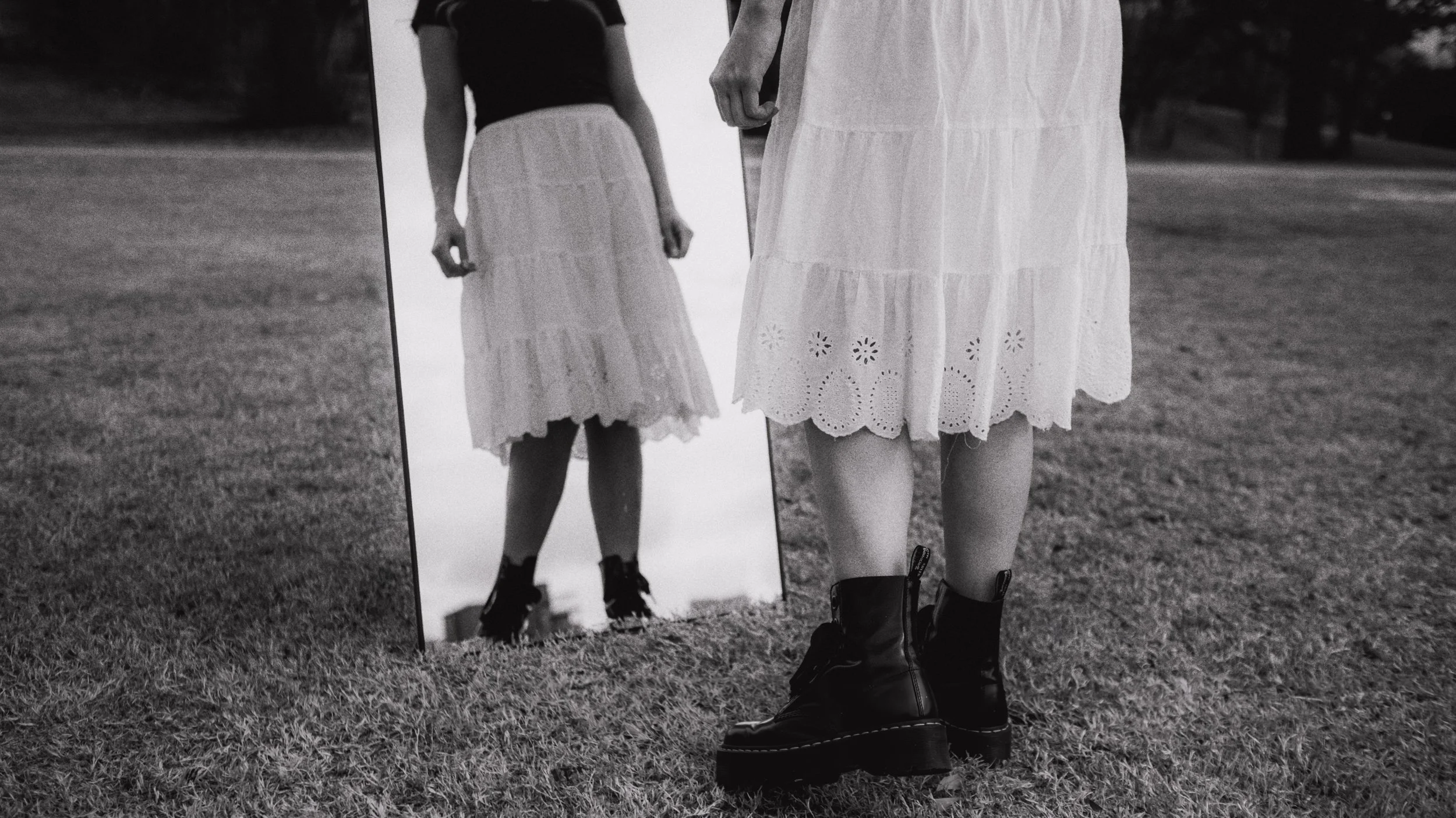 Close-up of a person's legs and lower body, wearing black combat boots and a white embroidered skirt, looking into a mirror reflecting their image, on grass in an outdoor setting.