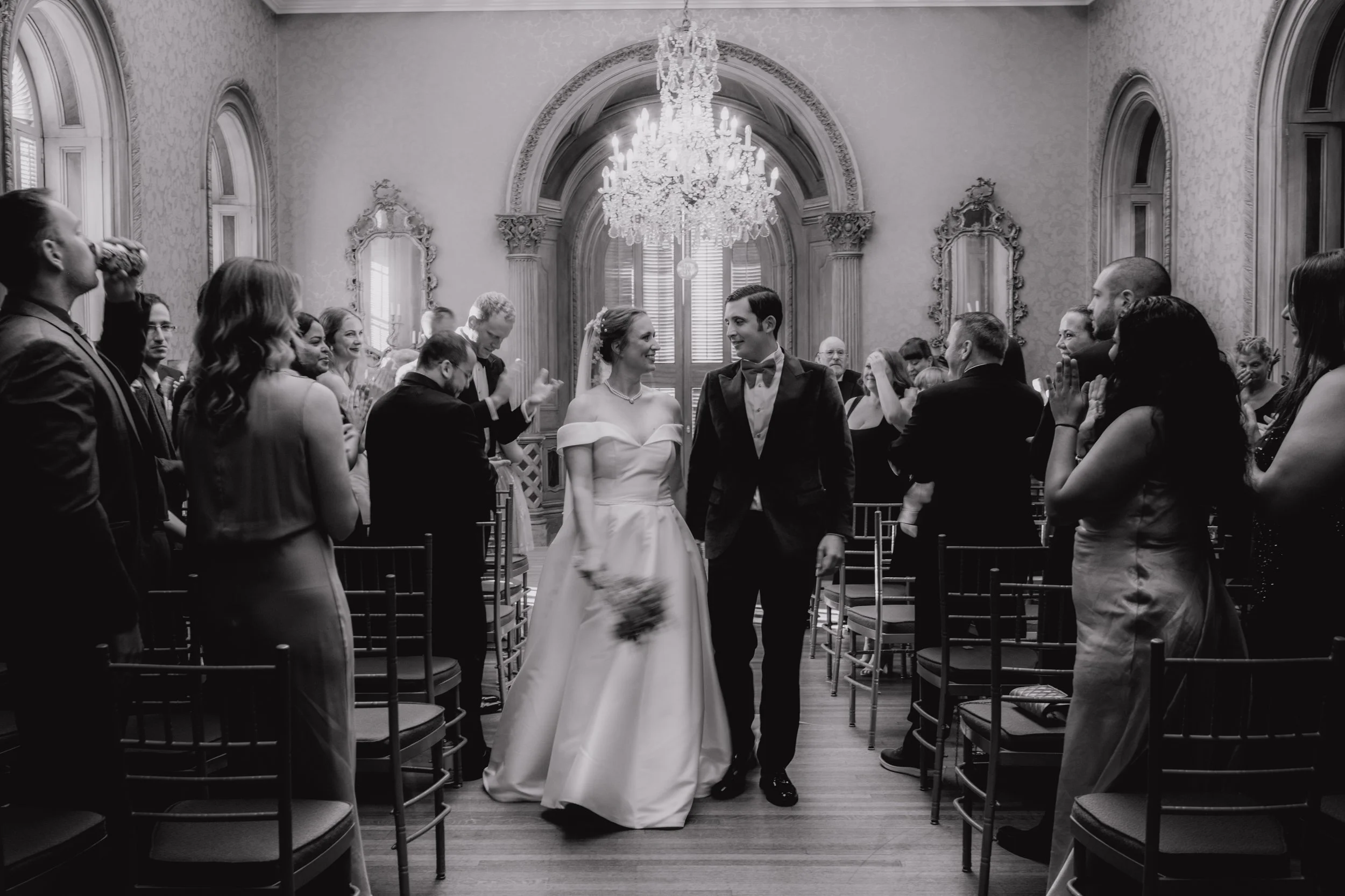 Black and white photo of a bride and groom walking down the aisle in a decorated wedding venue, surrounded by standing guests.