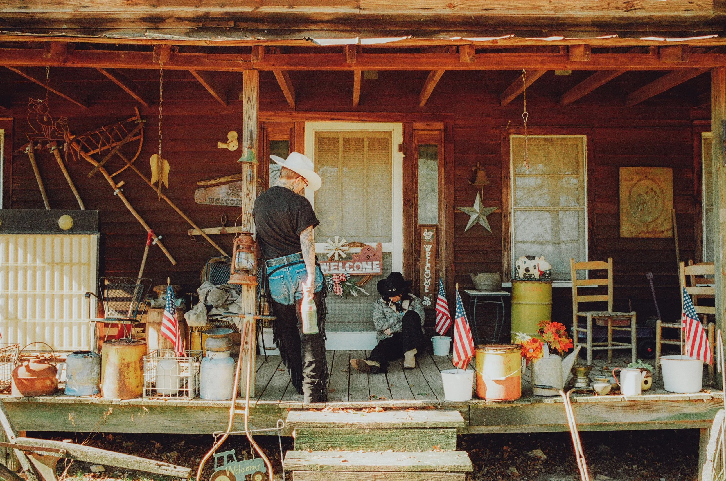 Front porch of a rustic wooden house decorated with American flags, a welcome sign, and farm tools. Two women in cowboy hats sit and stand, one holding a water bottle, on the porch.