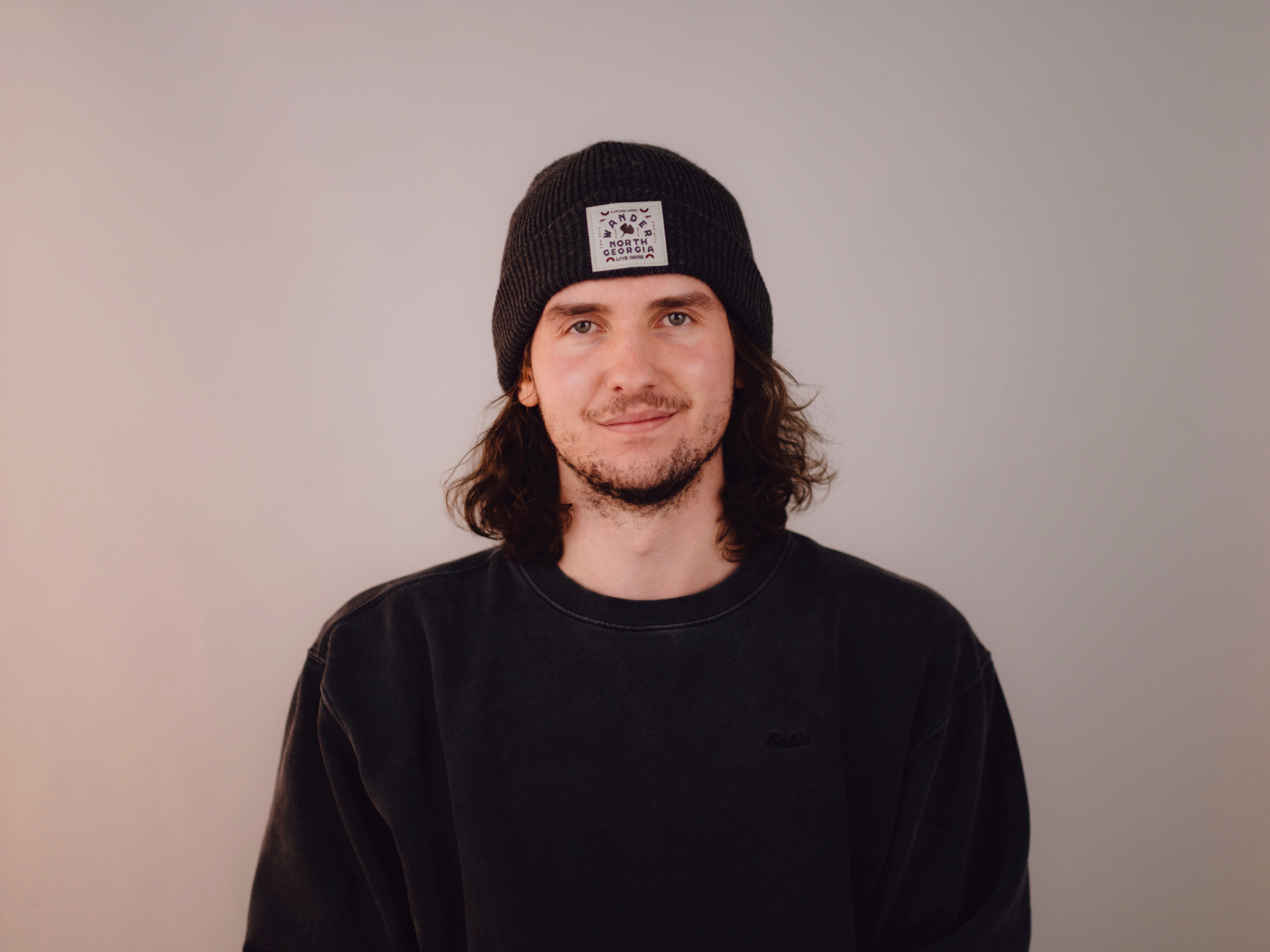 A young man with long brown hair, a slight beard, wearing a black knit beanie and a dark sweatshirt, standing in front of a plain light-colored background.