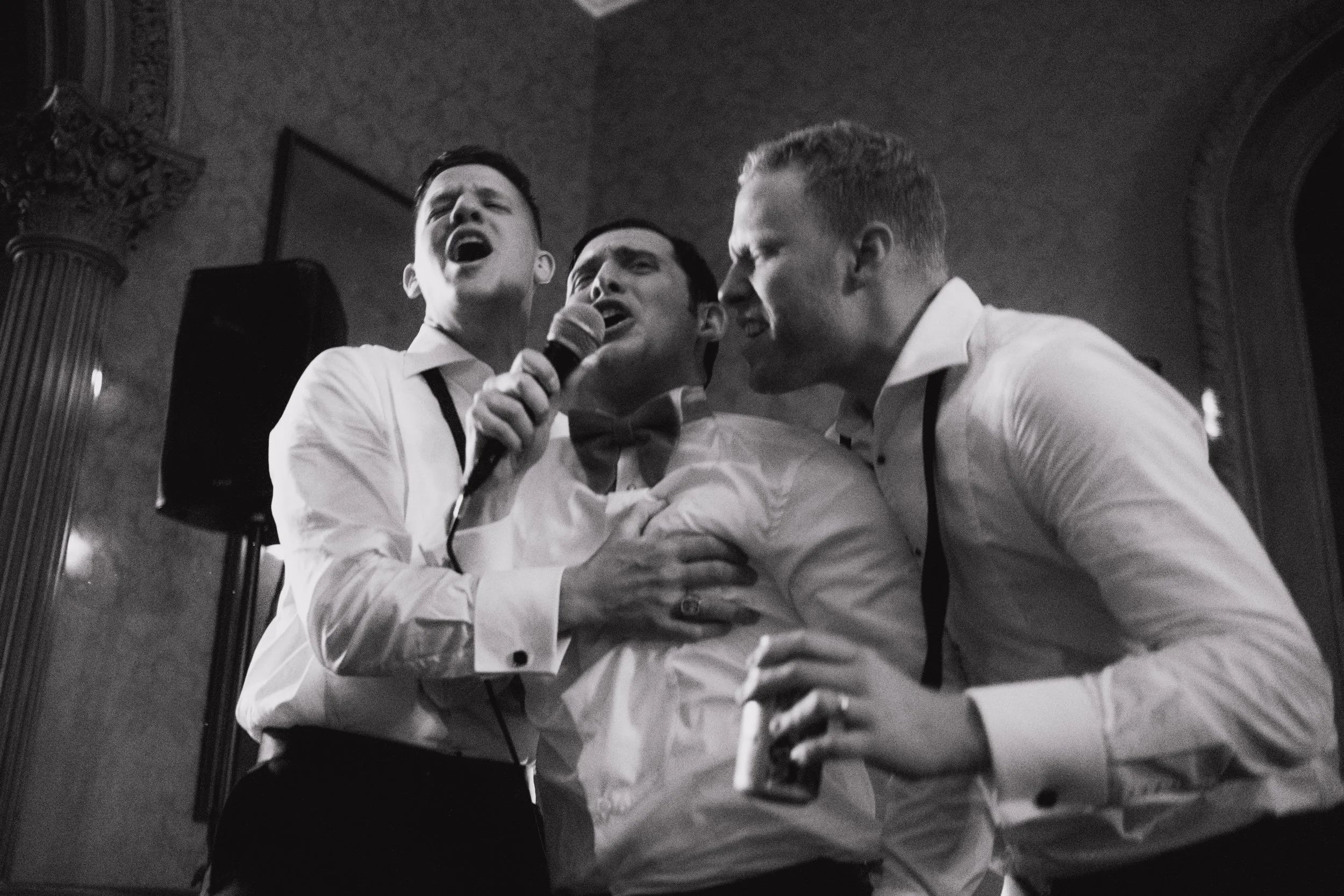Three men in formal attire singing passionately, one holding a microphone, in a dimly lit room with ornate decor.