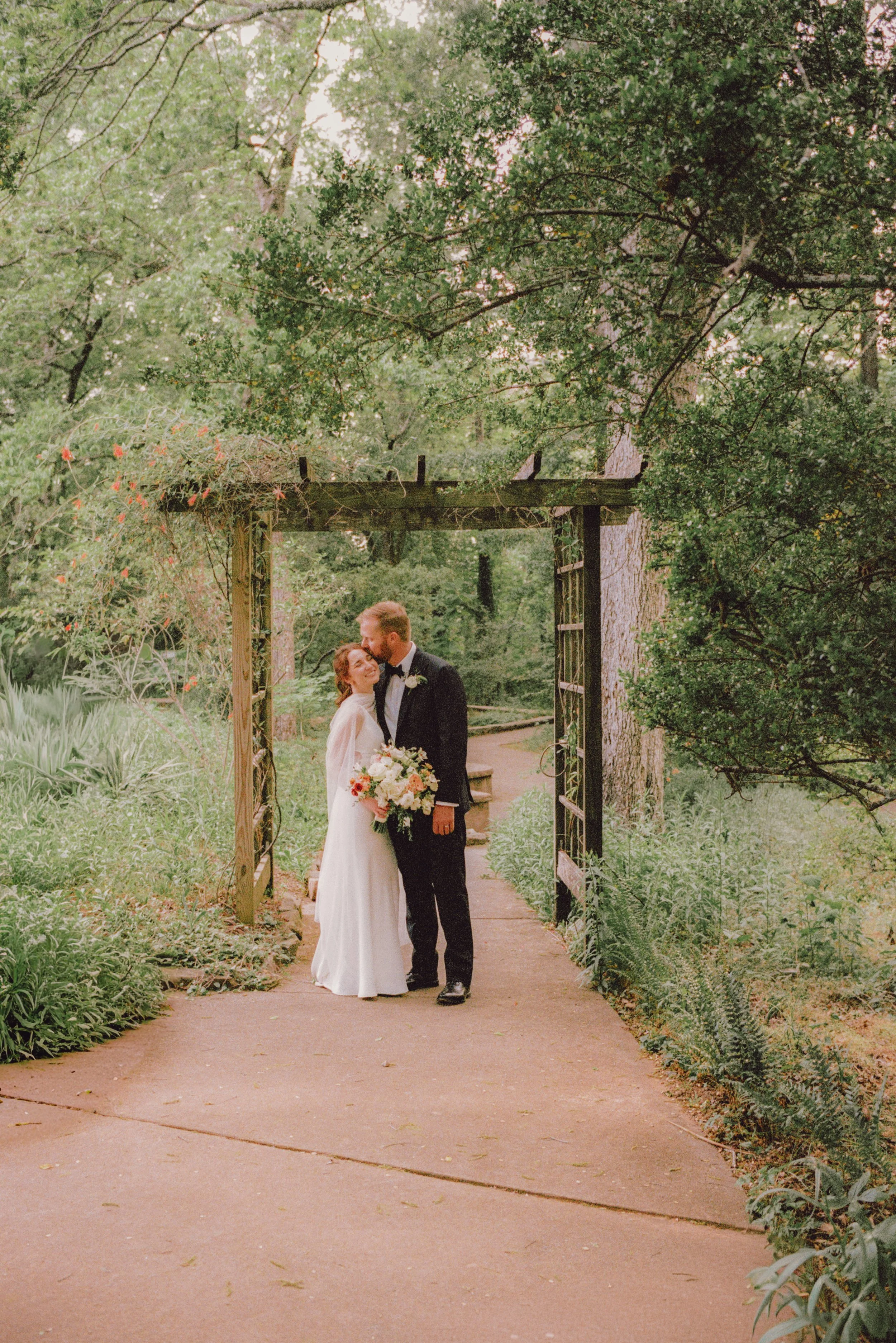 Bride and groom kissing beneath a wooden garden arbor on a tree-lined path at the Museum of Arts and Sciences in Macon