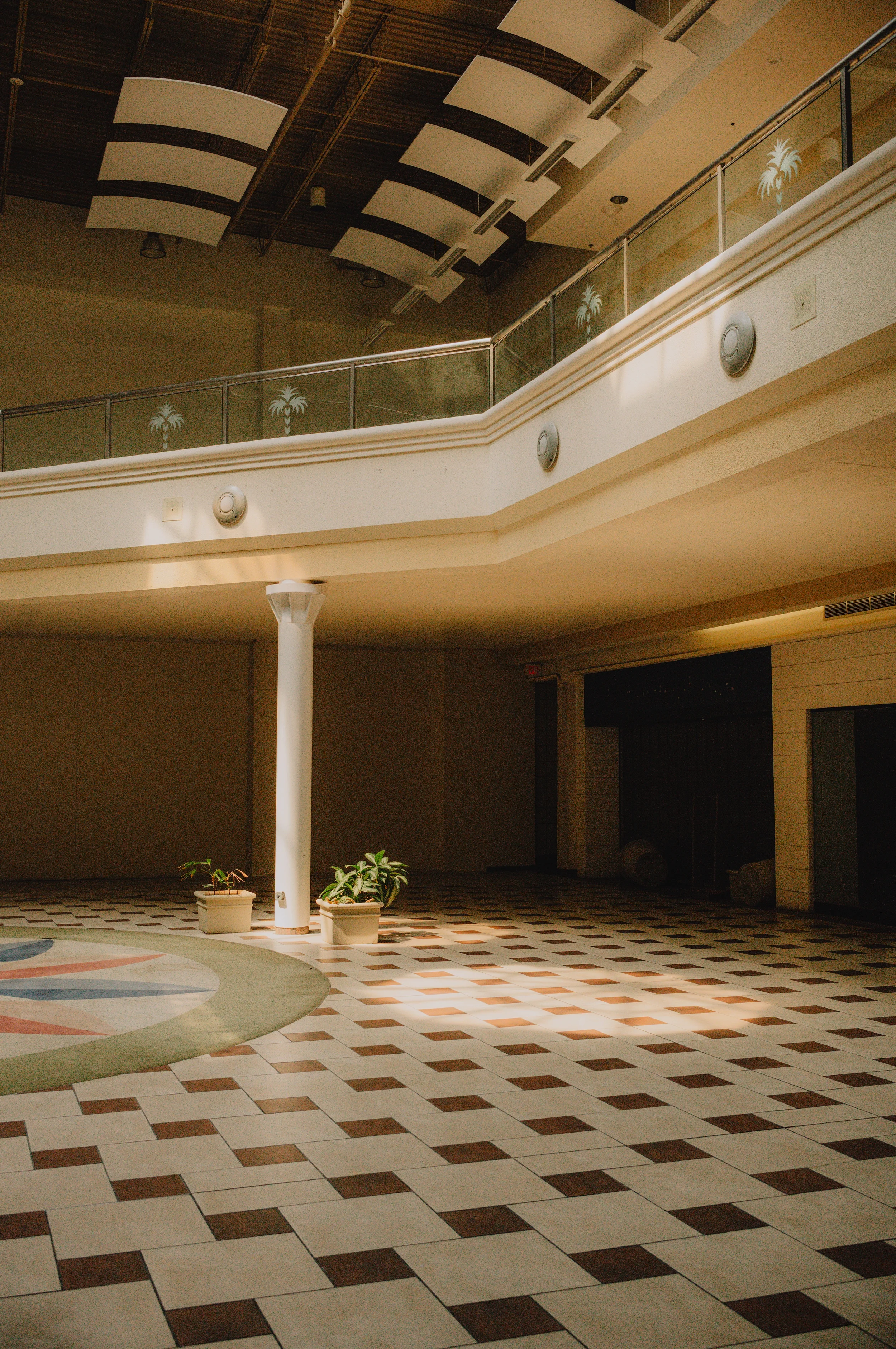 Empty indoor space with a tiled floor, potted plants, a white column, and a balcony with a glass railing decorated with palm tree decals. The ceiling has acoustic panels and industrial lighting.