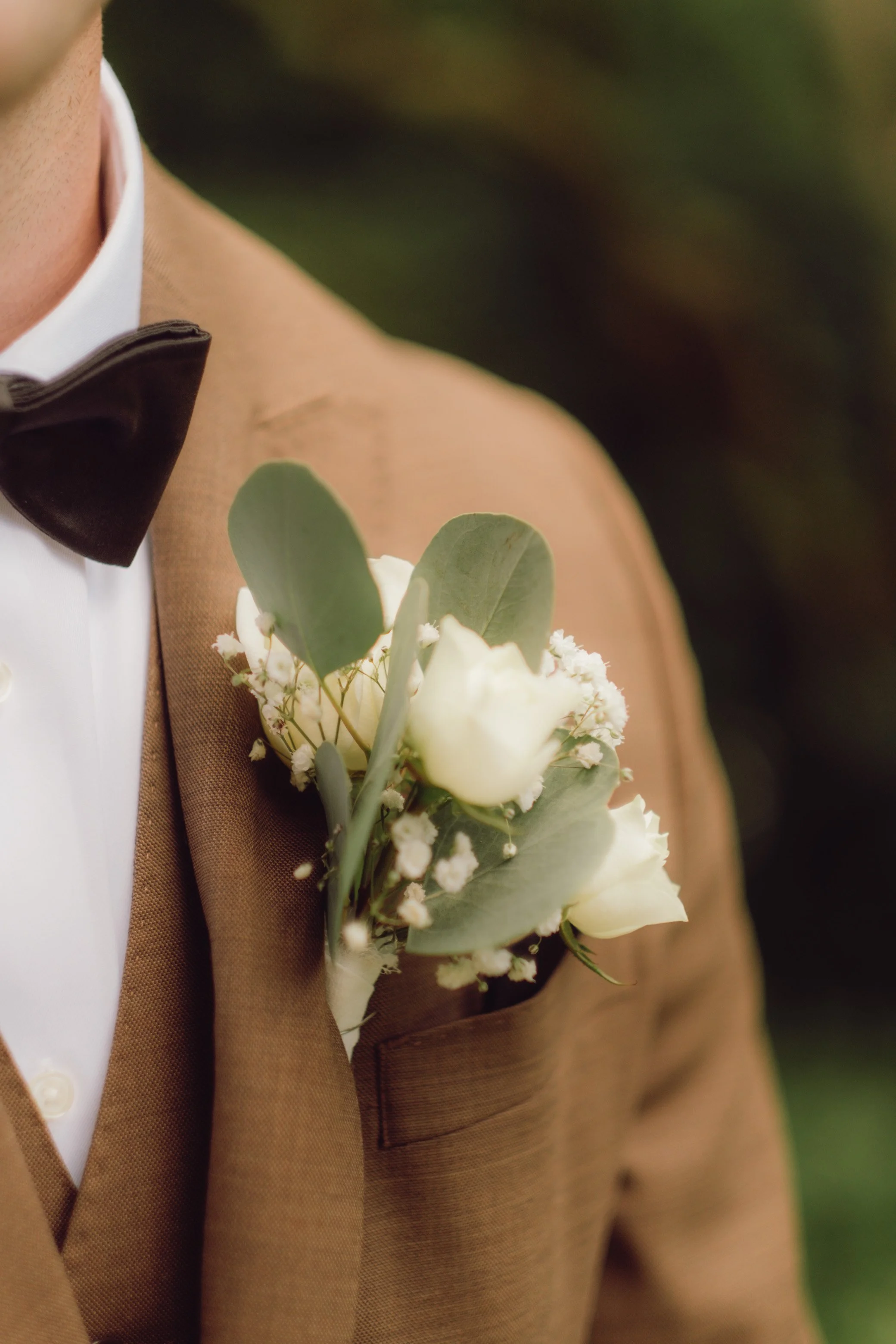 Close-up of a person wearing a brown suit, black bow tie, and white shirt with a boutonniere of white roses, baby's breath, and eucalyptus leaves pinned to the lapel.
