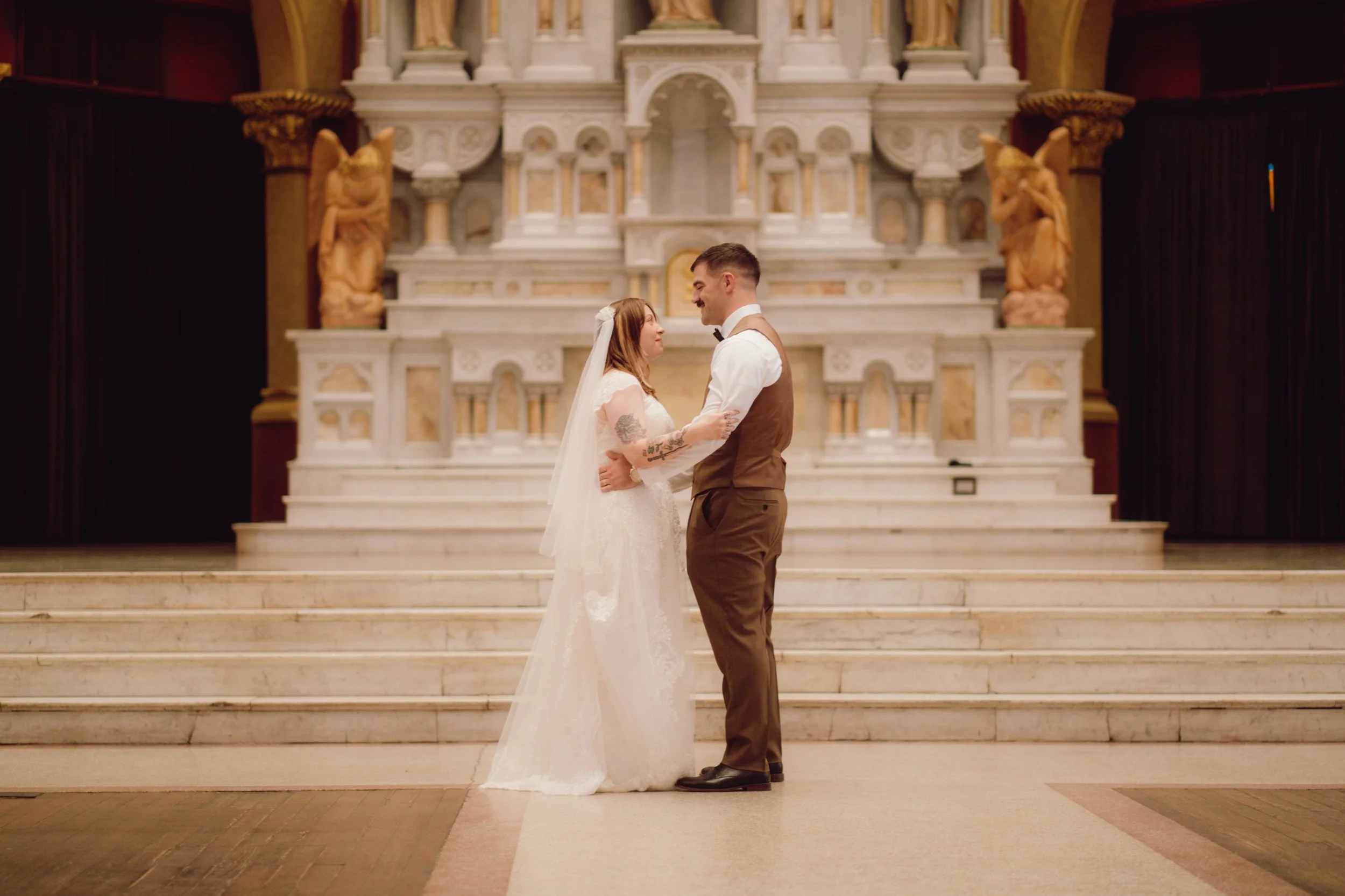 A bride and groom facing each other on wedding day, holding each other in a church with a marble altar in the background.