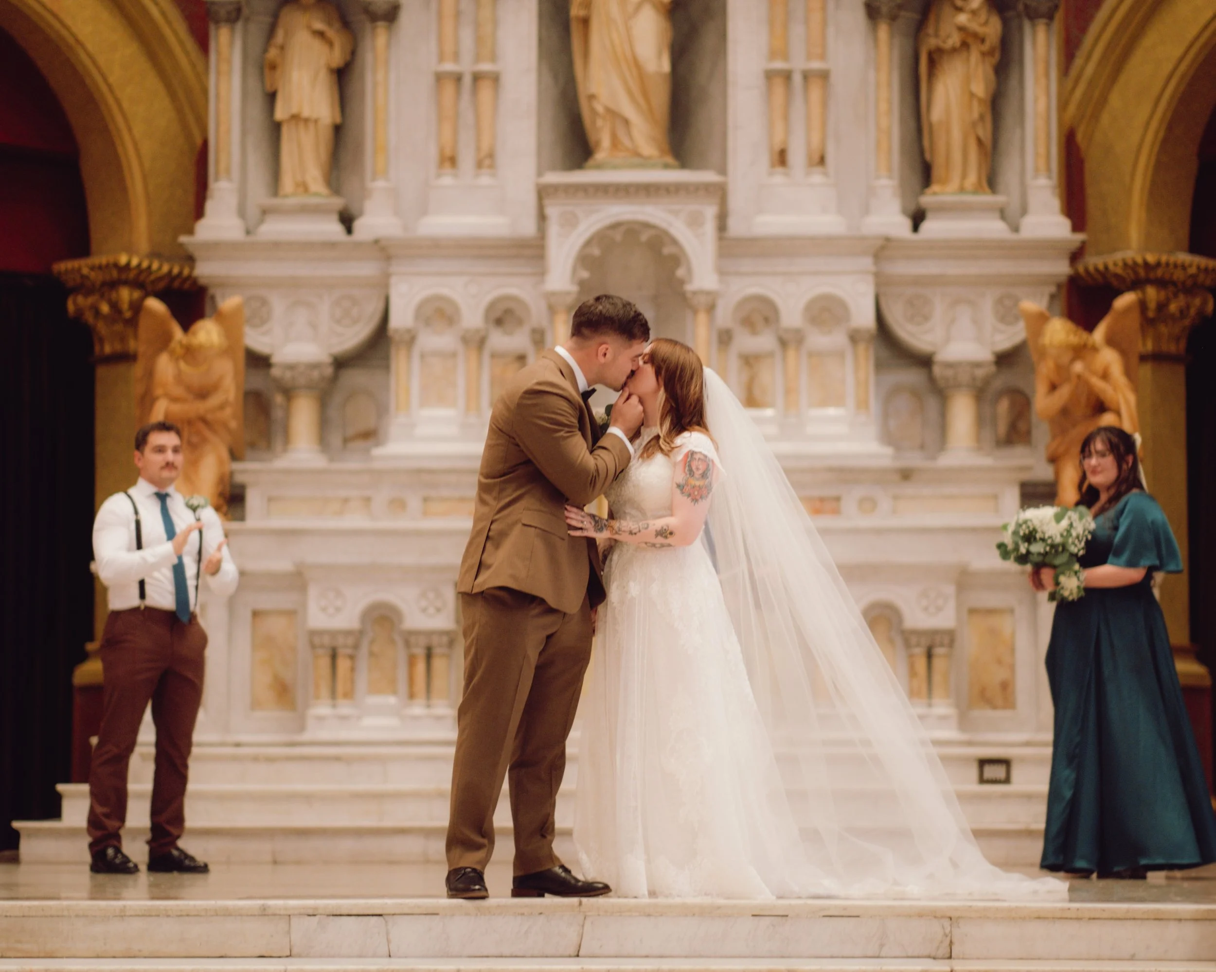 A couple getting married, sharing a kiss during their wedding ceremony in a church. The bride is wearing a white wedding dress with a long veil, and the groom is in a brown suit. Two people stand on either side, one holding a bouquet of flowers.