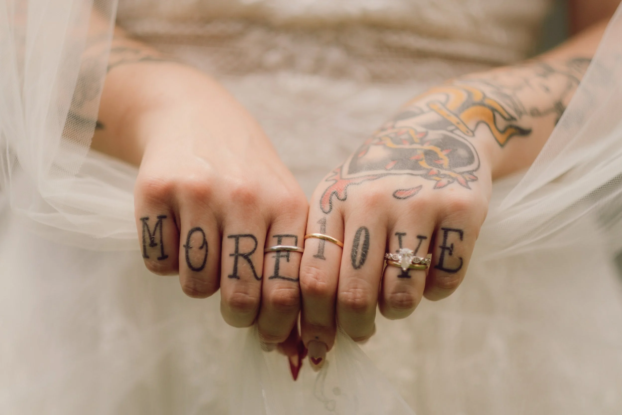 Two tattooed hands with rings, one with the word 'MORE' and the other with 'LOVE' tattooed on the fingers, extended forward with a light-colored fabric in the background.