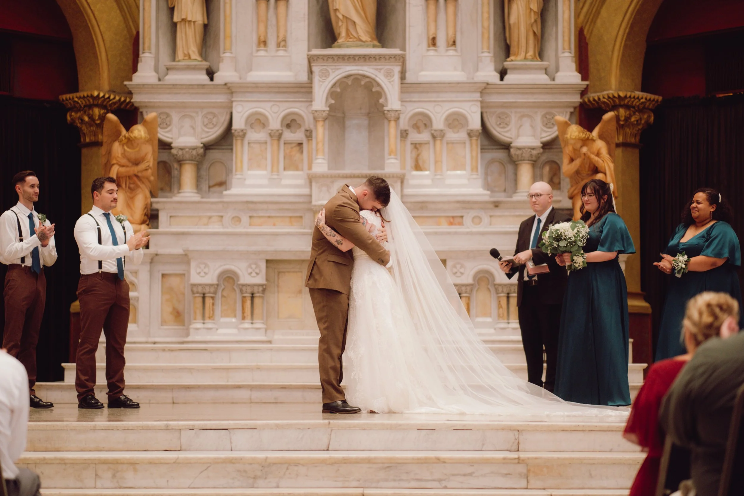 A wedding ceremony taking place on marble stairs inside a church, with a couple embracing and kissing at the altar, surrounded by bridesmaids, groomsmen, and officiants, all in formal attire.