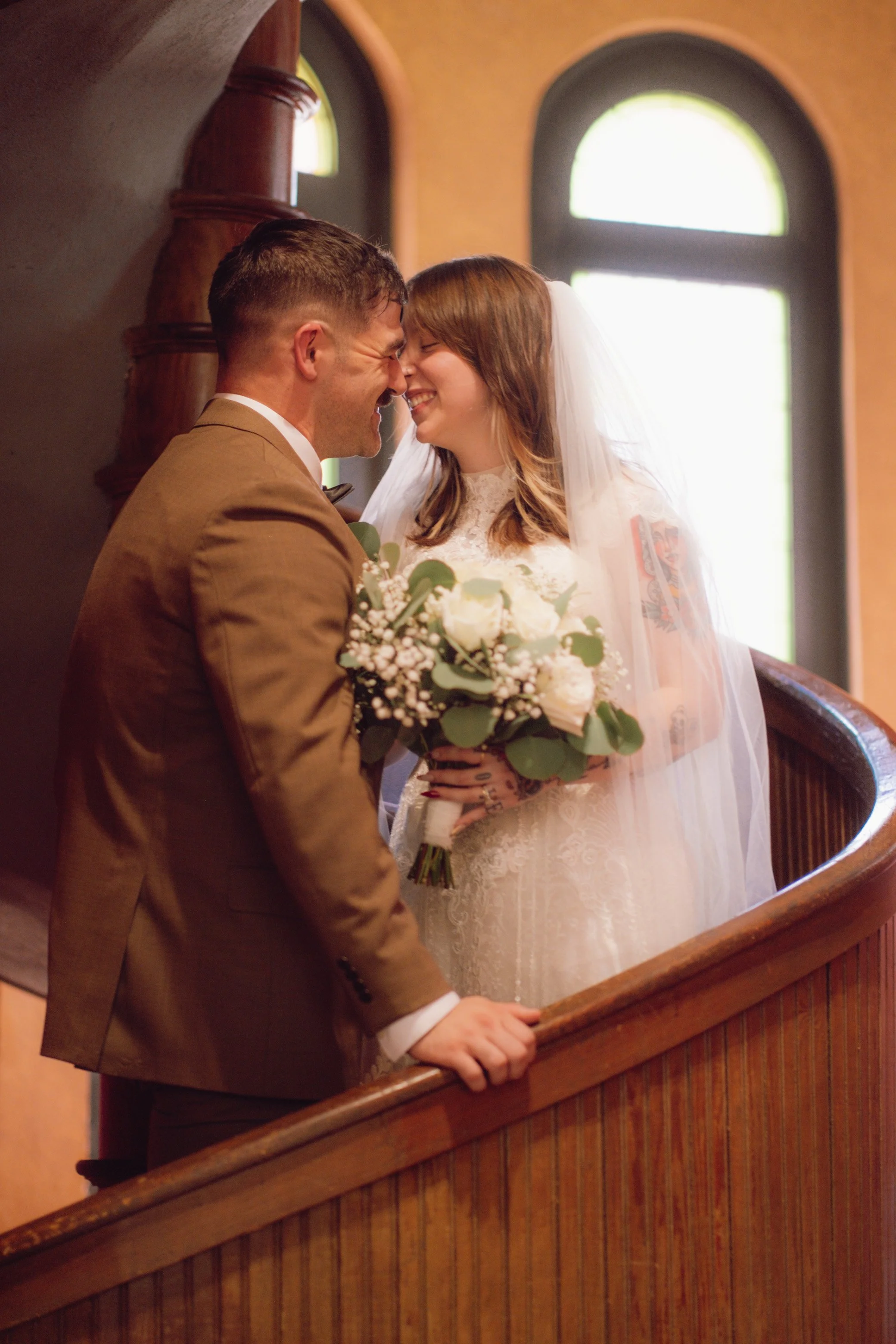 A newlywed couple sharing a joyful moment with their foreheads touching, inside a church. The groom is dressed in a brown suit and the bride in a white lace wedding gown with a veil, holding a bouquet of white roses and greenery.