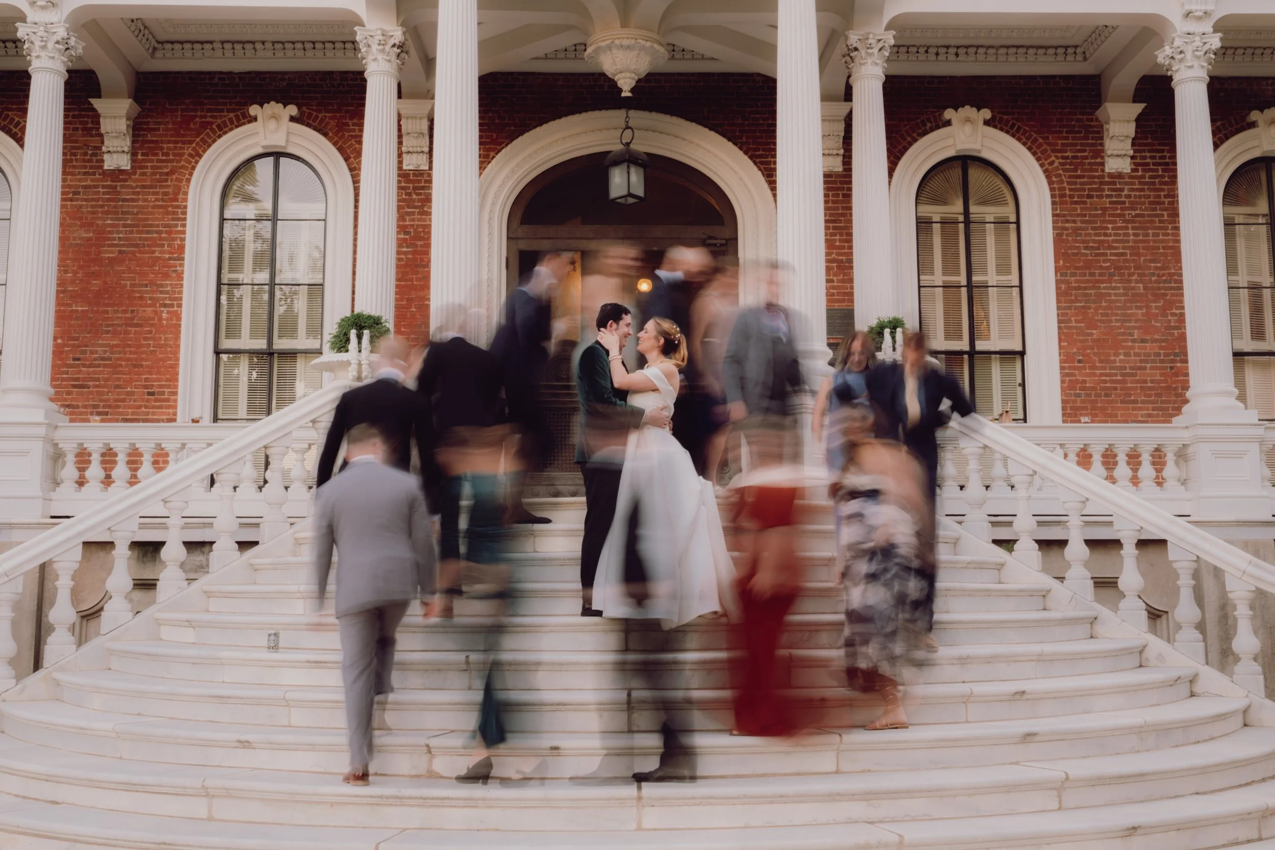 A bride and groom sharing a kiss on the steps of a grand brick mansion, surrounded by moving guests in formal attire at a wedding.