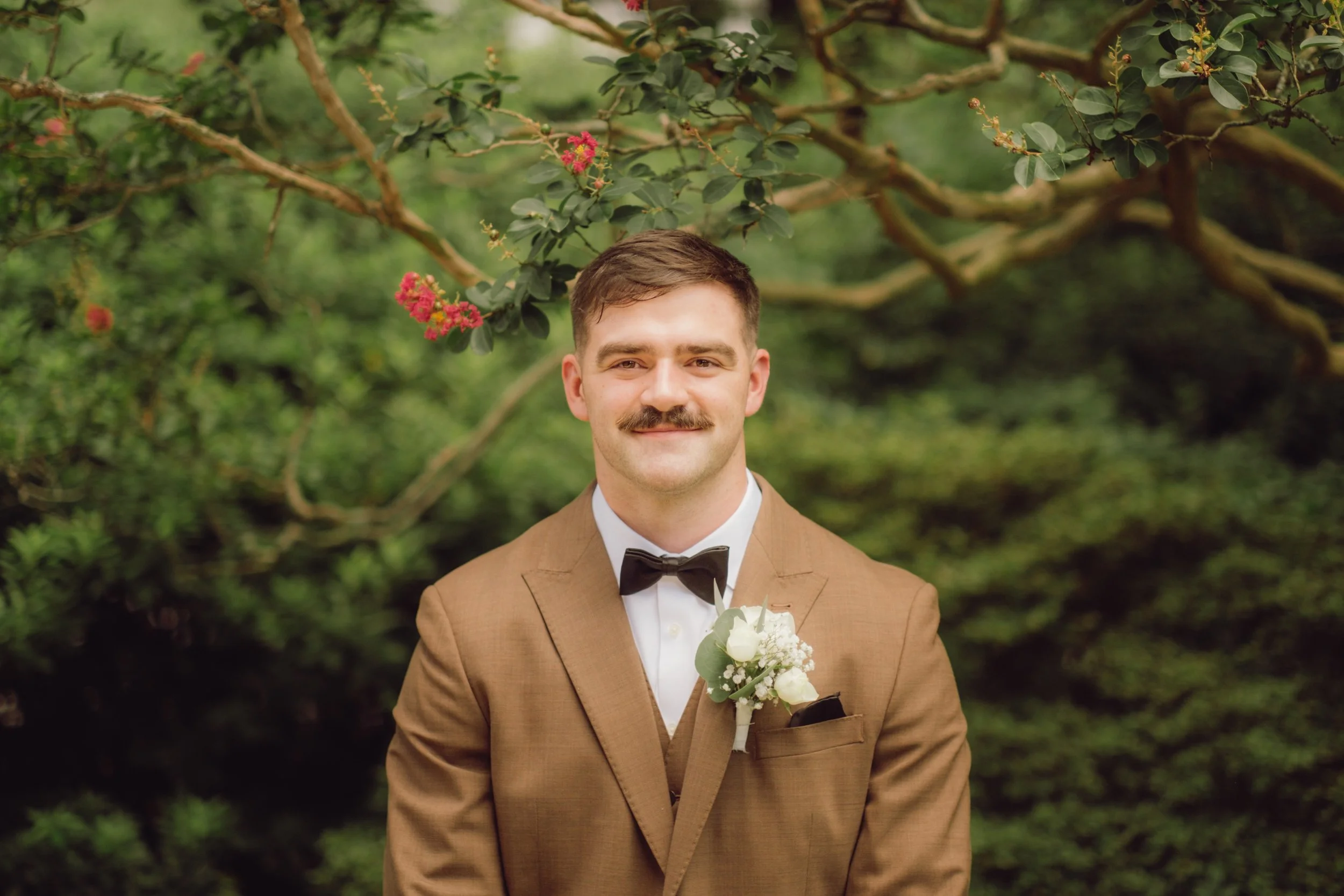 A man with brown hair, a mustache, and a slight smile wearing a brown suit with a white shirt and black bow tie, standing outdoors in front of green foliage and a branch with pink flowers, with a white boutonniere on his lapel.