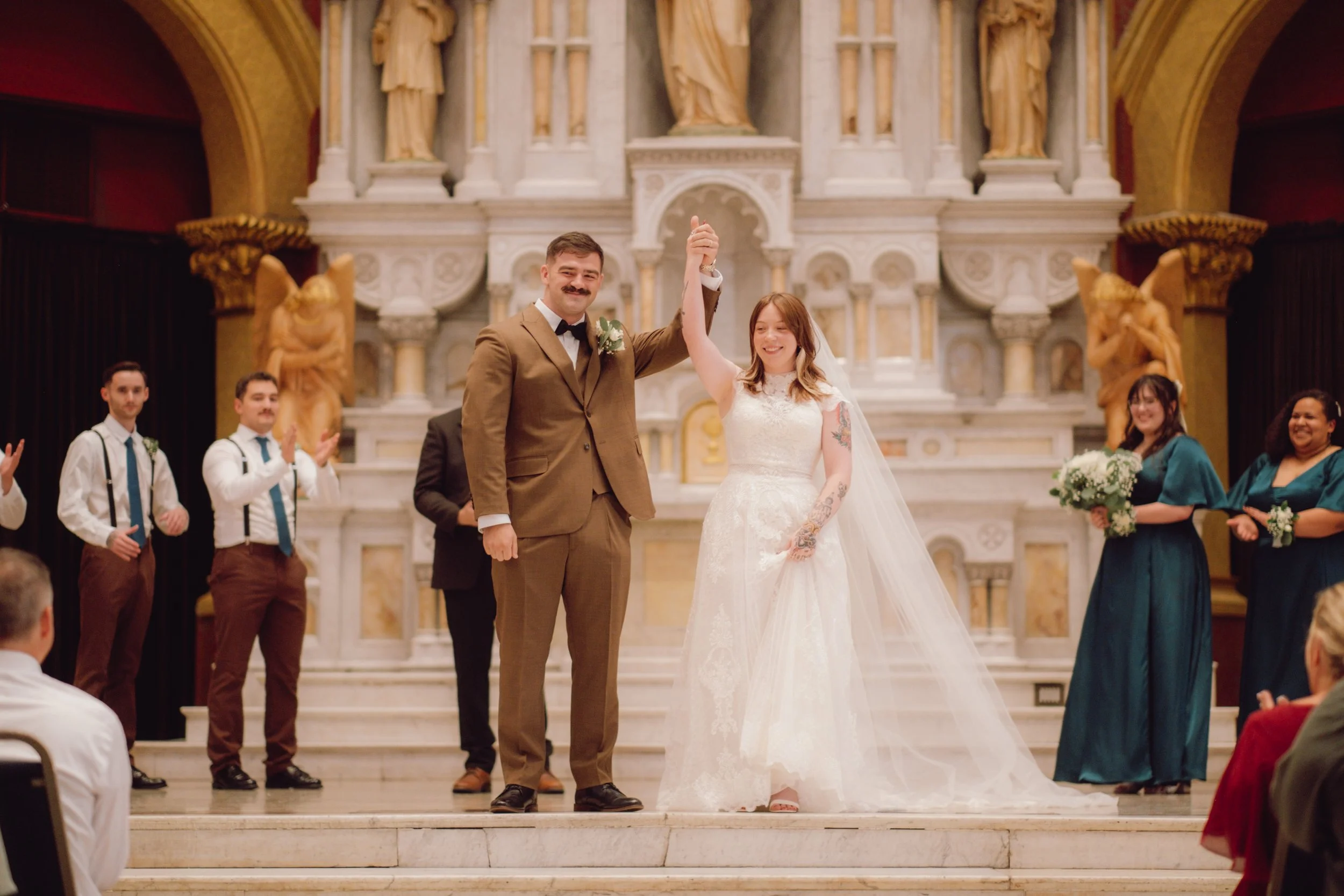 A newly married couple on their wedding day, standing on a church altar, celebrating with friends and family. The groom is wearing a brown suit and bowtie, and the bride is in a white lace wedding dress with a long veil. They are holding hands up in