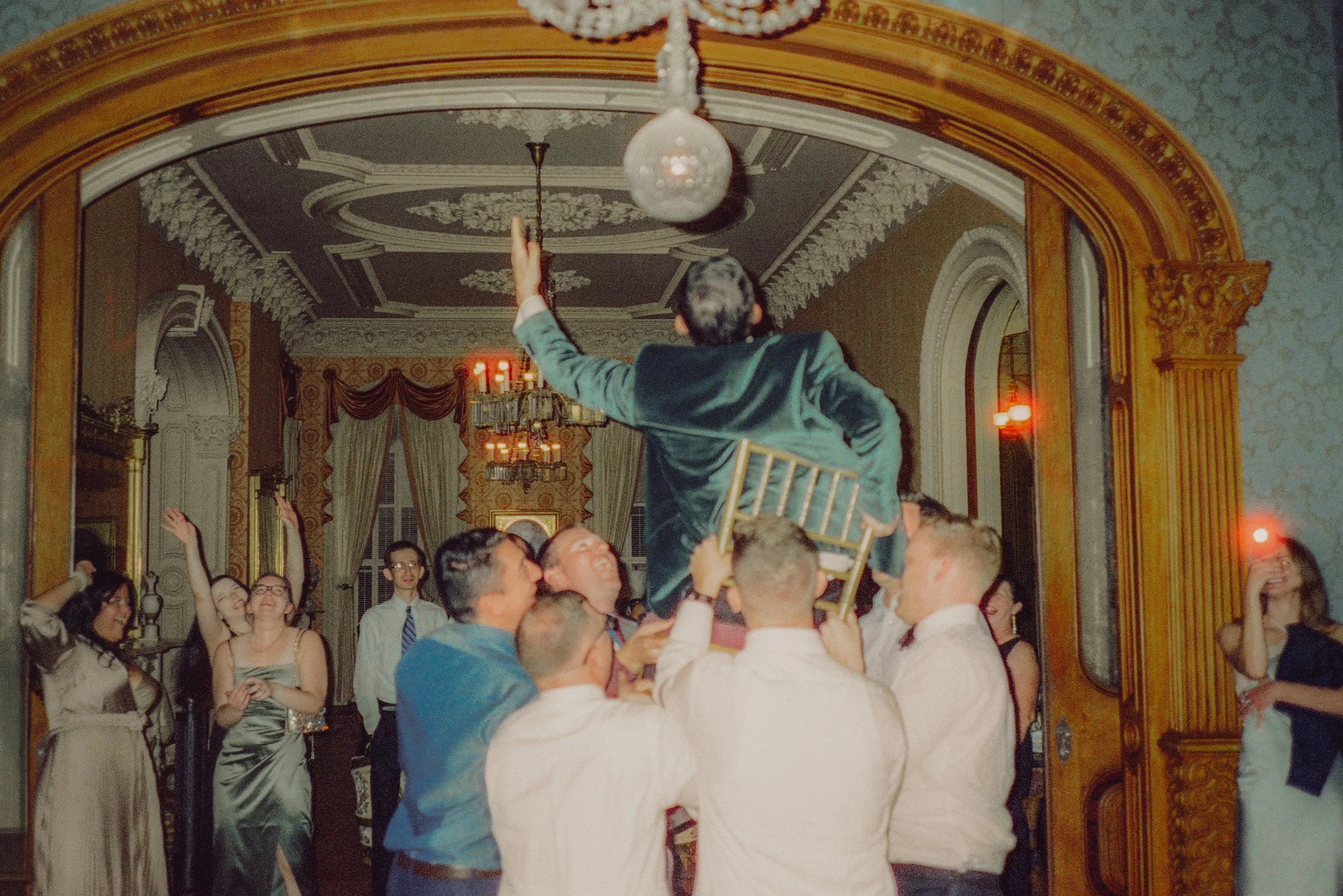 Groom lifted up in a chair by guests during the reception in the Hay House ballroom