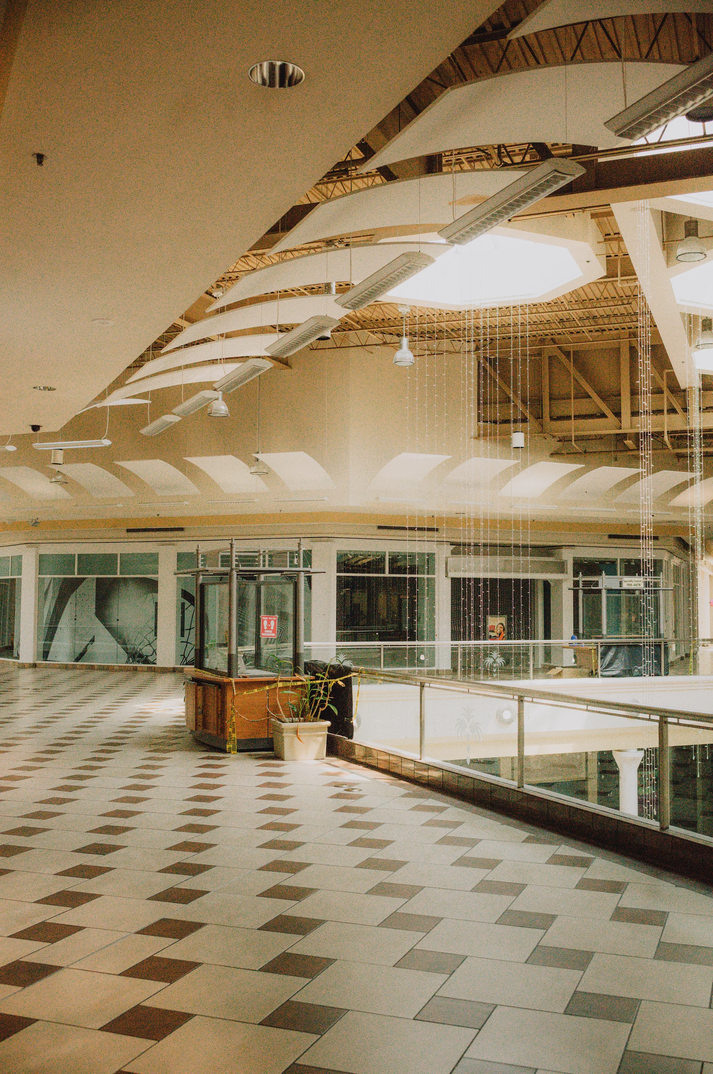 Interior view of a shopping mall with an open ceiling structure, large skylight, hanging light fixtures, and a tiled floor with a diamond pattern.