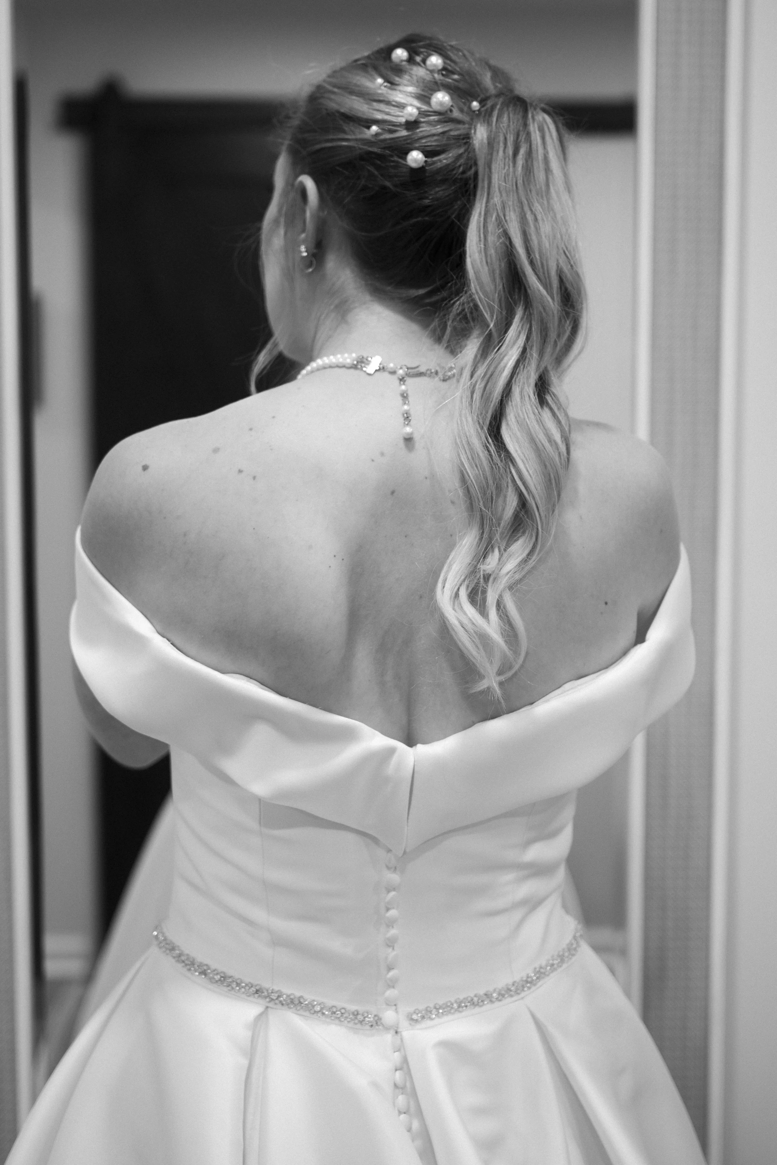 A woman in a wedding dress with an off-the-shoulder neckline, jewelry, and styled hair, seen from behind, standing in an elevator.