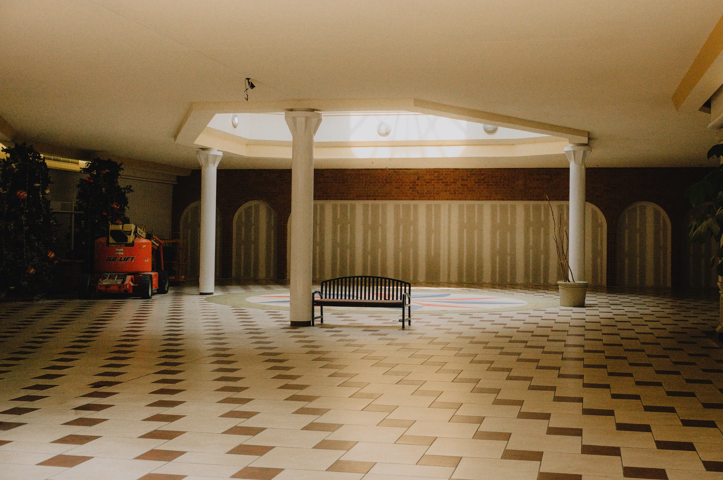 Empty indoor space with patterned tile flooring, a large skylight, potted plants, a bench, and some equipment on the left side.