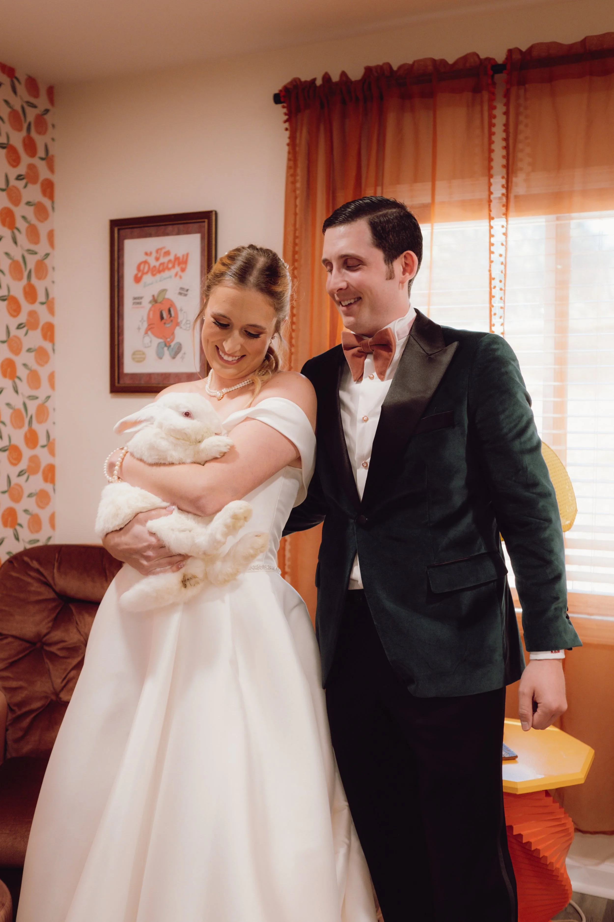 A newlywed couple smiling and holding a white rabbit indoors, with the bride dressed in a white wedding dress and the groom in a black tuxedo and bowtie, in a room with orange curtains and retro decor.