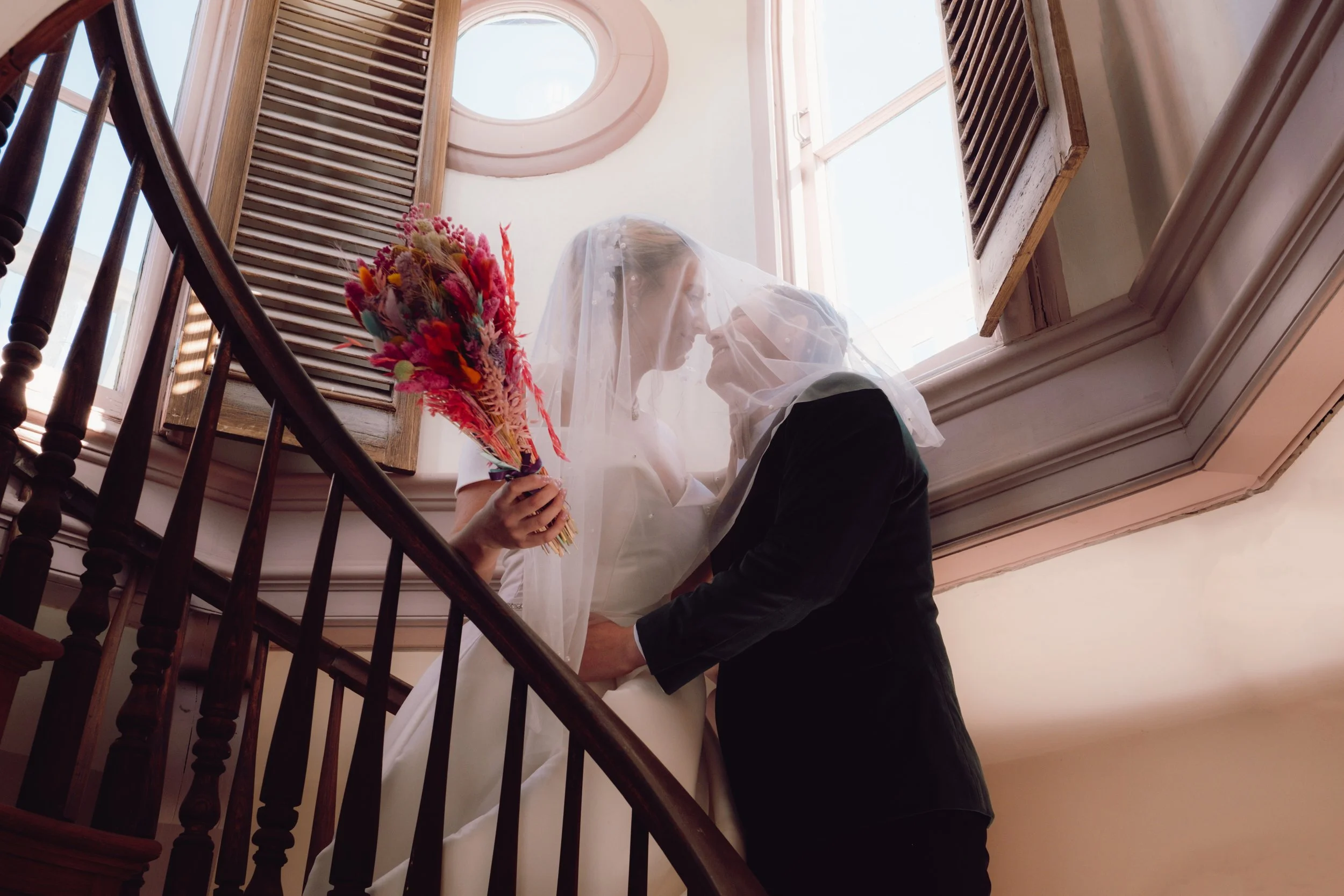 A bride and groom standing on a staircase, with the bride holding a colorful bouquet and both leaning towards each other with a veil over their faces. The scene is set indoors near a window with wooden shutters.