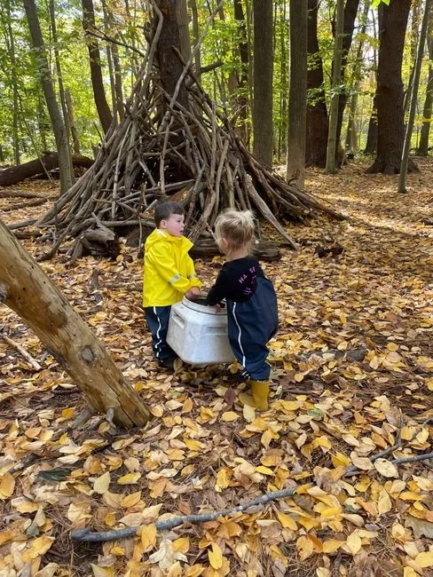 Two children in forest classroom, working together. The ground is covered with fallen leaves, and there is a small teepee made of sticks behind them.
