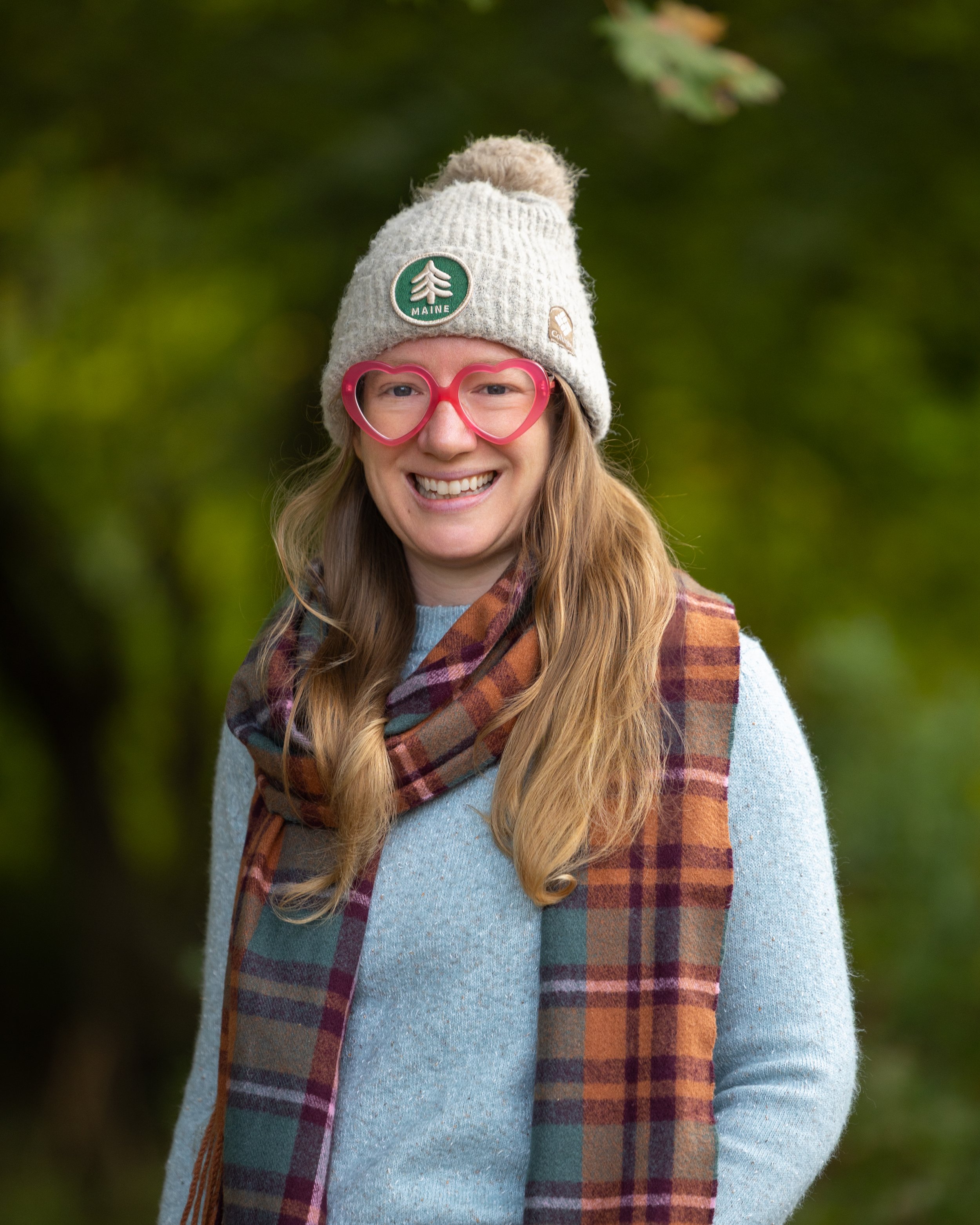 A woman with long blonde hair smiling outdoors, wearing a beige knit hat with patches, pink heart-shaped glasses, a light blue sweater, and a multicolored plaid scarf.