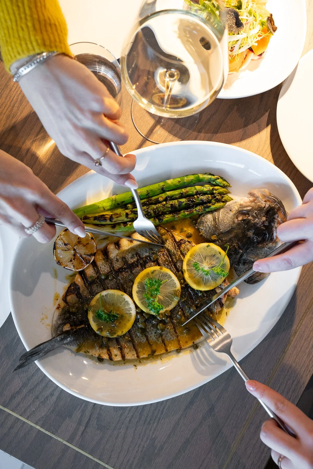 Grilled whole fish with charred lemon and asparagus as two diners serve portions from the shared plate