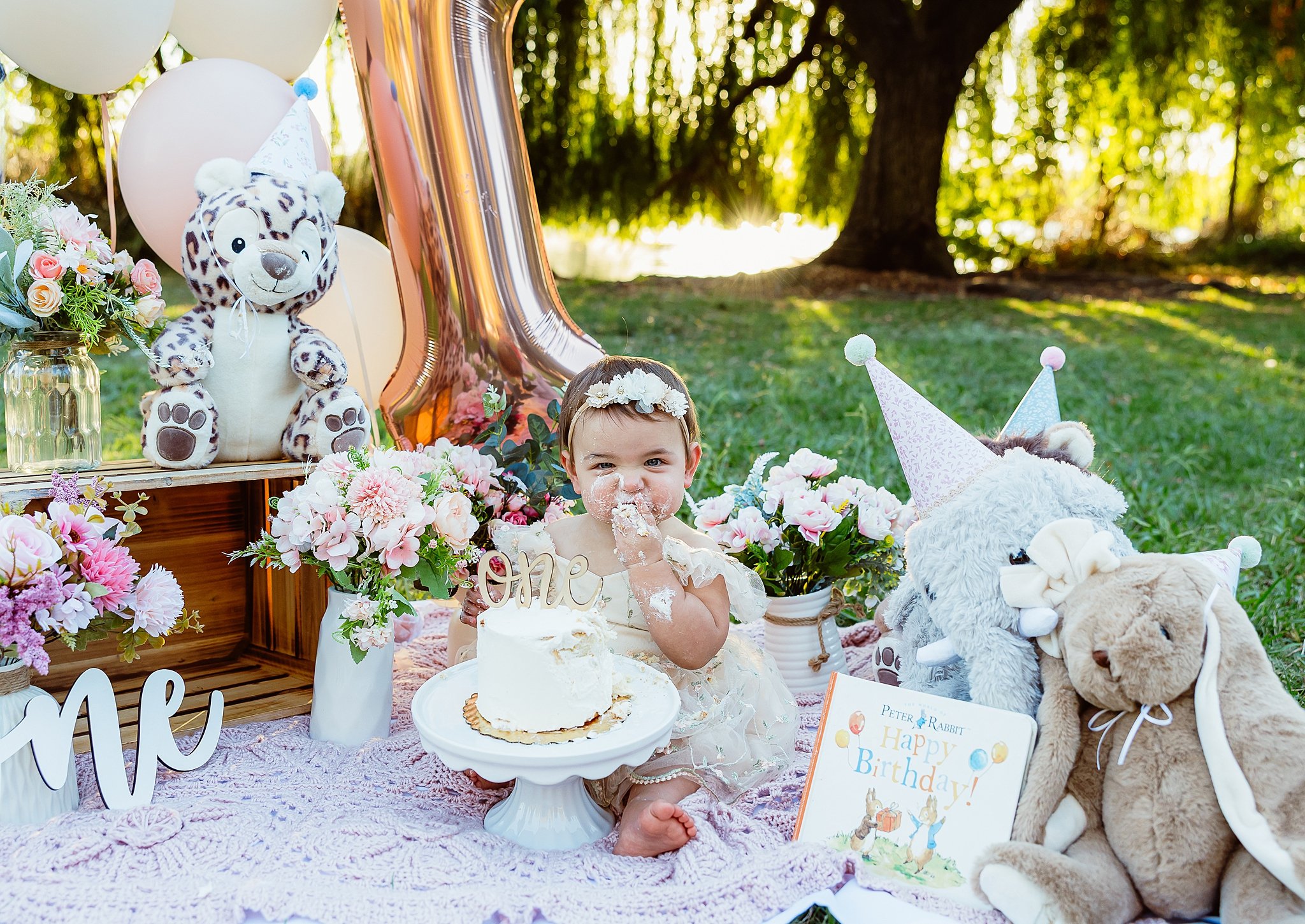 First birthday cake smash session with baby girl surrounded by balloons, flowers, and stuffed animals during an outdoor milestone session in Elk Grove, CA