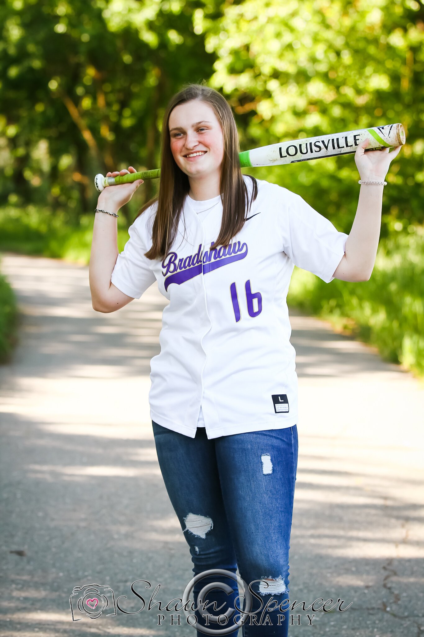  senior girl wearing jeans and softball jersey holding bat over back of head on shoulders 