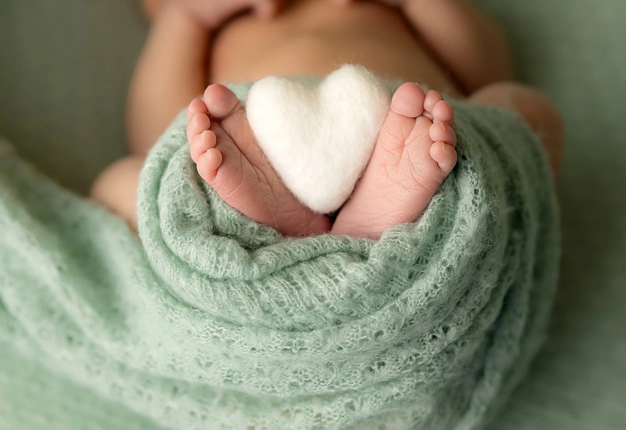 Close-up detail of newborn baby feet wrapped in soft green fabric holding a small white heart during an Elk Grove newborn photography session.