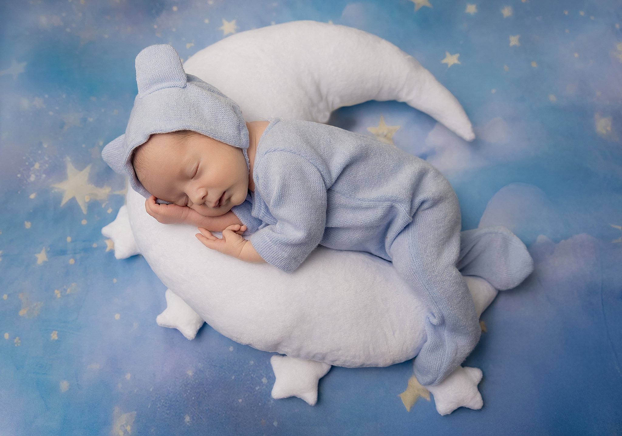 Sleeping newborn baby in a soft blue outfit posed on a crescent moon prop with a starry backdrop in an Elk Grove home studio.