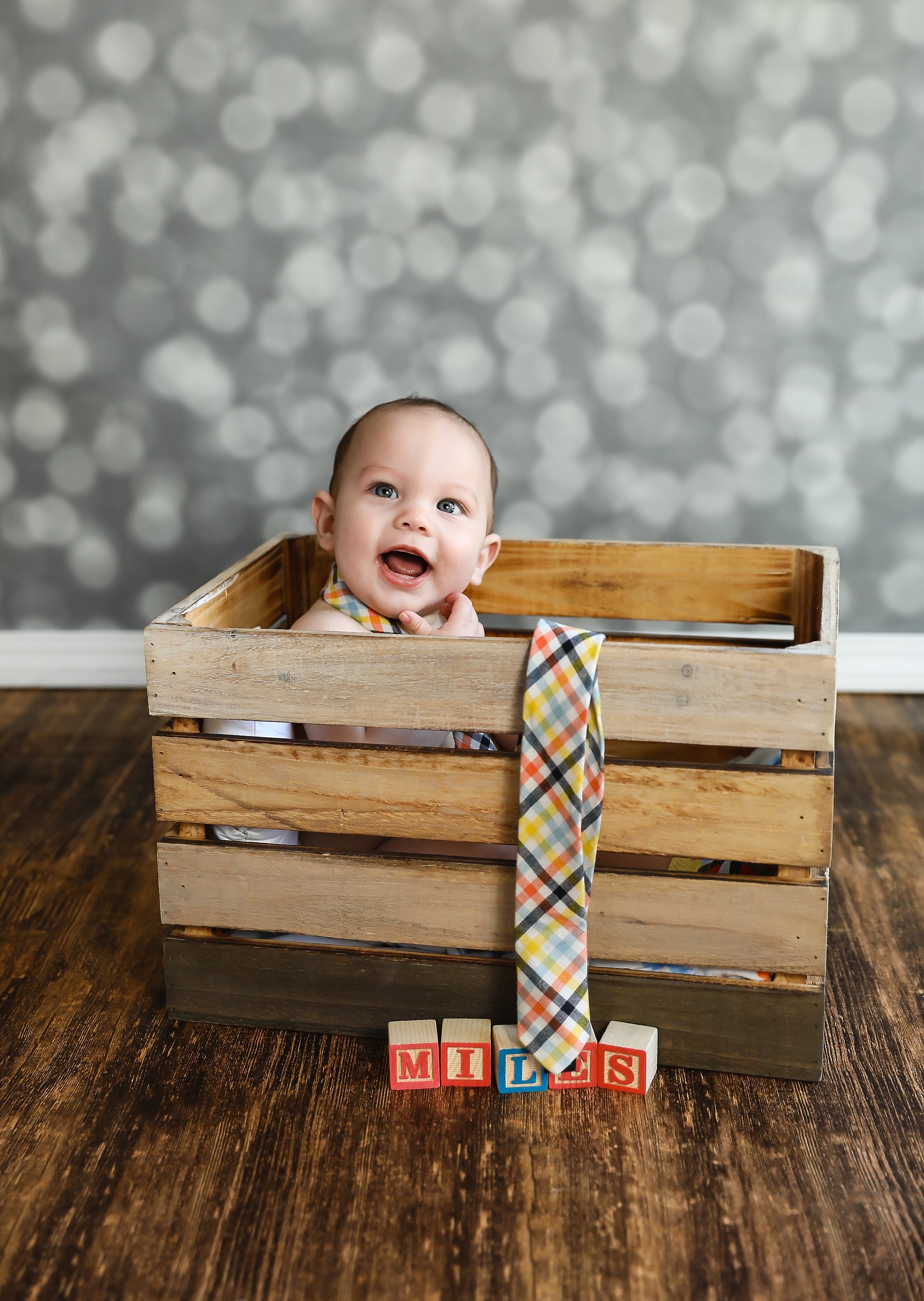 Smiling six-month-old baby boy sitting inside a wooden crate during a studio milestone portrait session in Elk Grove, California