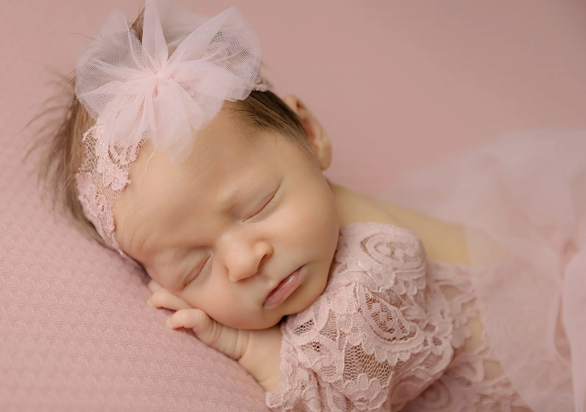 Close-up portrait of a sleeping newborn baby girl wearing a soft blush lace outfit and bow headband in an Elk Grove home studio.