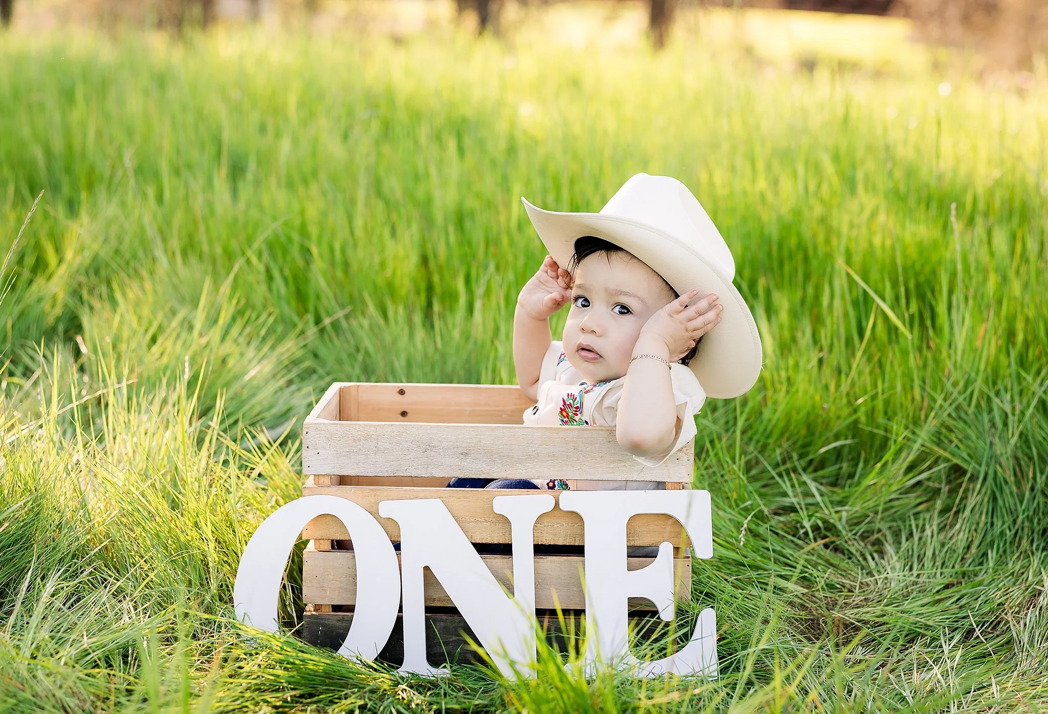 One-year-old boy wearing a cowboy hat while sitting in a wooden crate with "ONE" letters during an outdoor first birthday portrait session in Elk Grove, California.