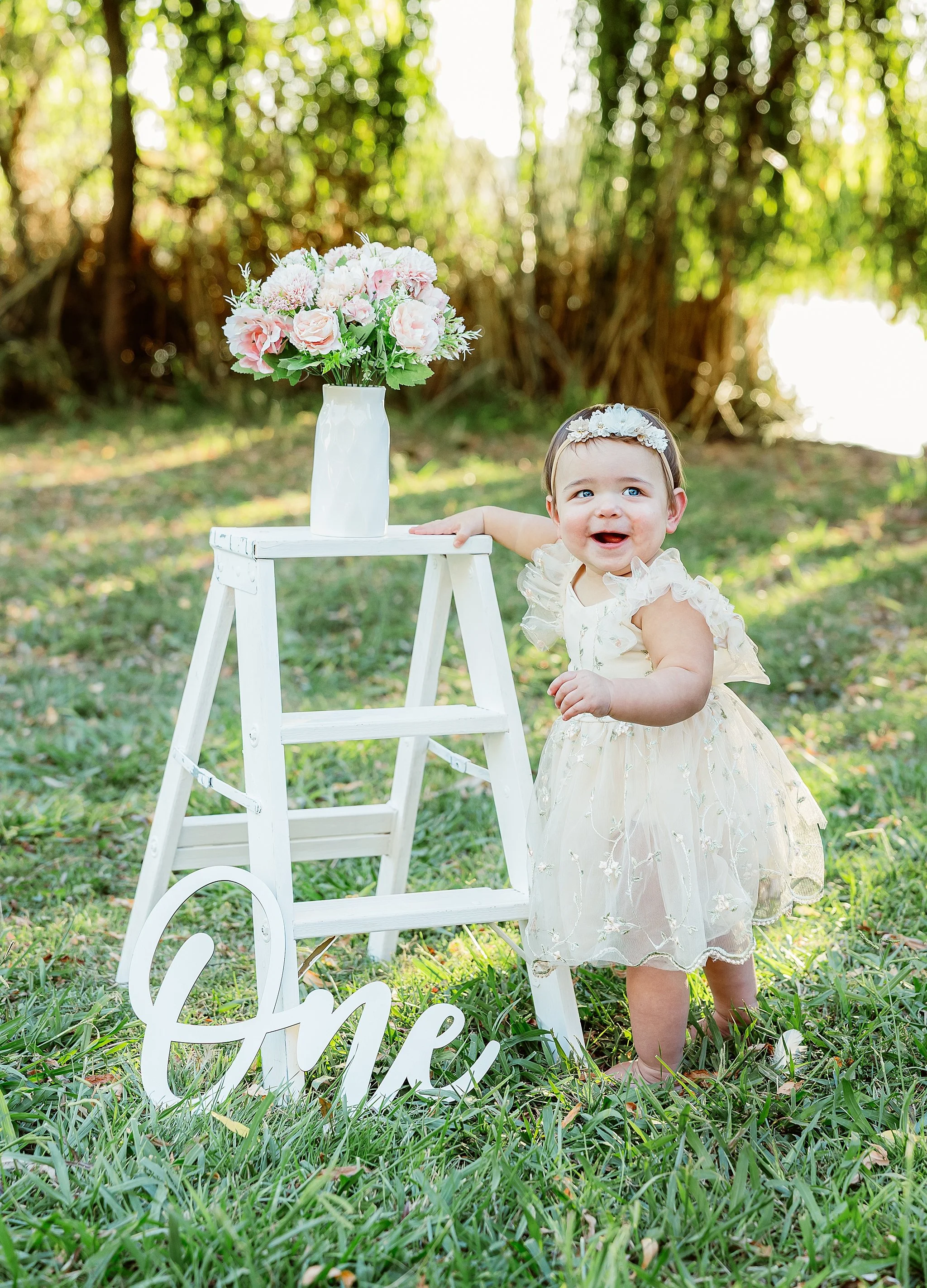 One-year-old baby girl standing beside a stool with florals during an outdoor milestone portrait session in Elk Grove