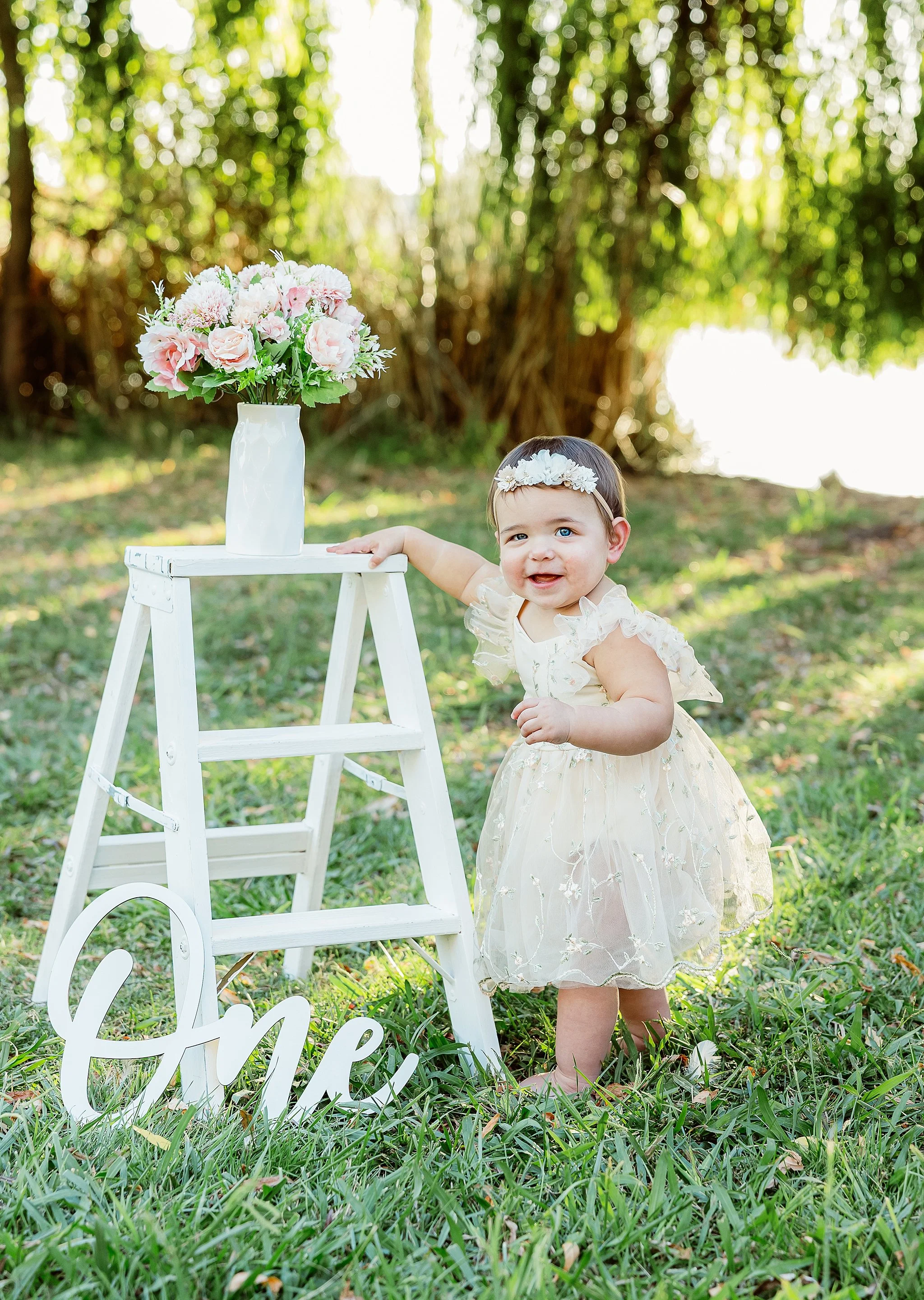 One-year-old girl standing beside a white ladder with pink flowers during an outdoor first birthday milestone portrait session in Elk Grove, California.
