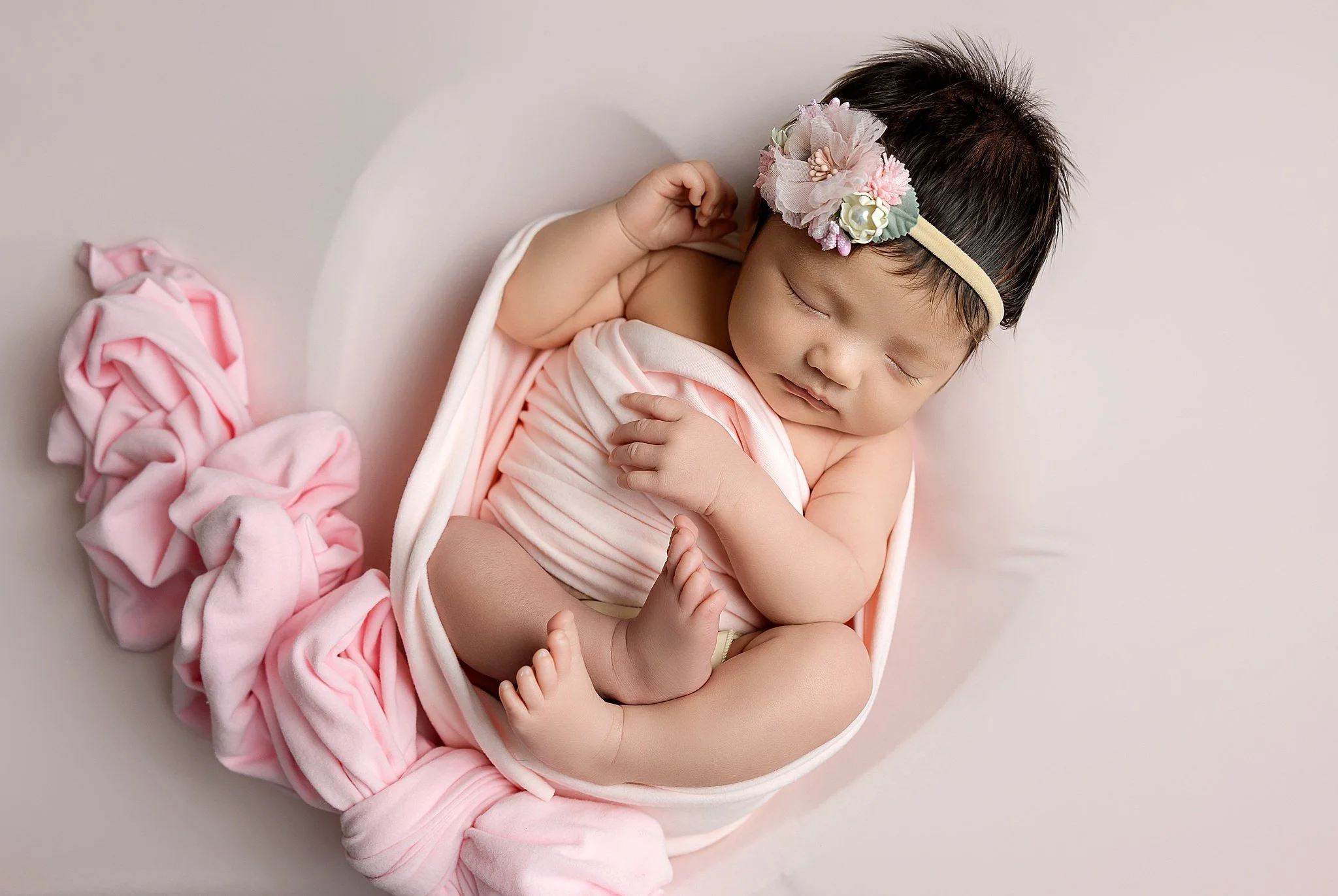 Newborn baby girl wrapped in soft pink fabric wearing a delicate floral headband photographed in an Elk Grove home studio.