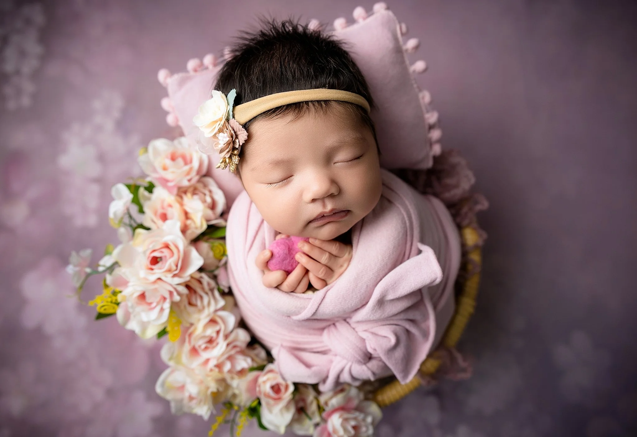 Sleeping newborn baby girl posed in a basket with soft pink roses and a pastel backdrop during a studio session in Elk Grove, California.