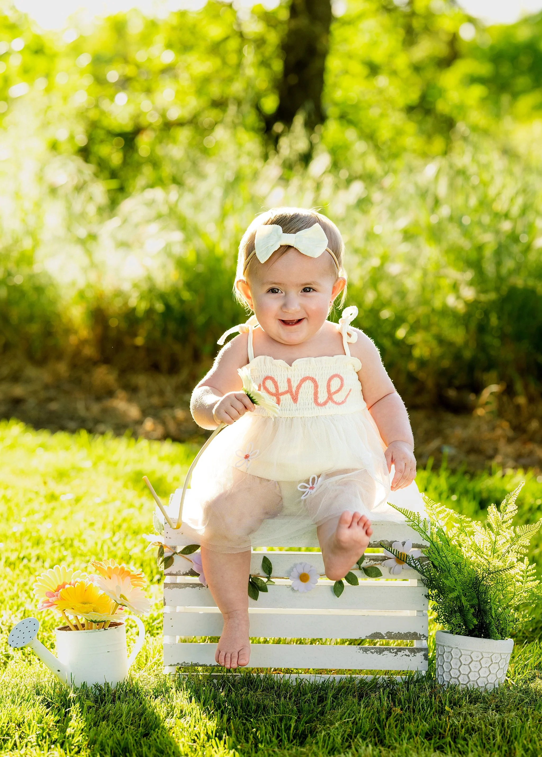 Smiling one-year-old girl sitting on a white crate surrounded by flowers during an outdoor first birthday milestone portrait session in Elk Grove, California
