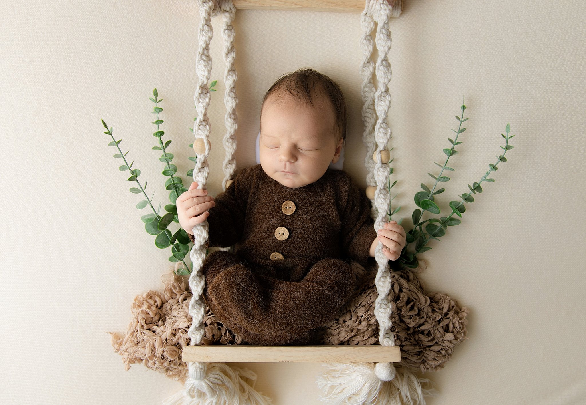 Newborn baby boy wearing a brown knit outfit posed on a decorative swing with greenery accents in an Elk Grove newborn studio.