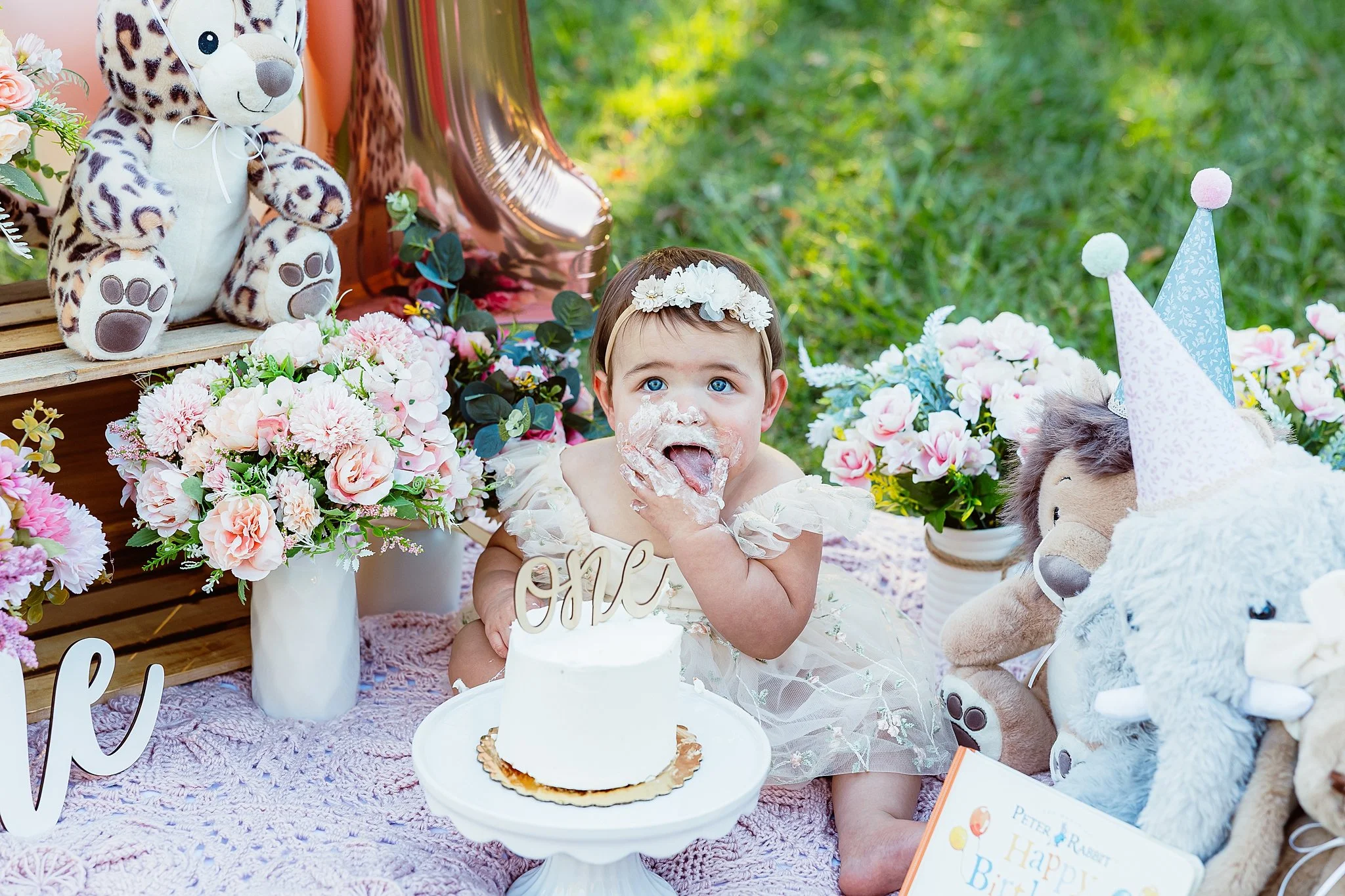 One-year-old girl enjoying her cake smash surrounded by flowers and stuffed animals during an outdoor first birthday photography session in Elk Grove, California.