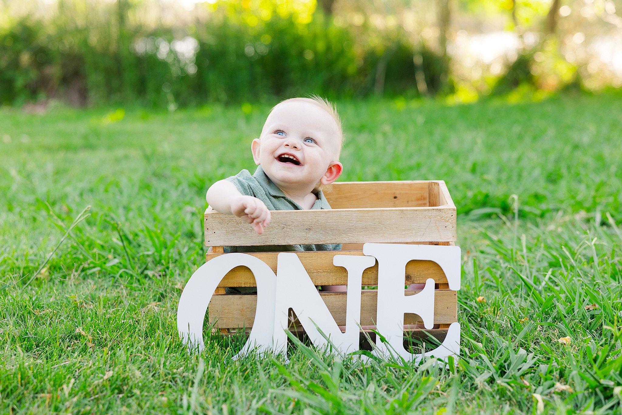 Happy one-year-old boy peeking out of a wooden crate behind large "ONE" letters during an outdoor first birthday portrait session in Elk Grove, California.